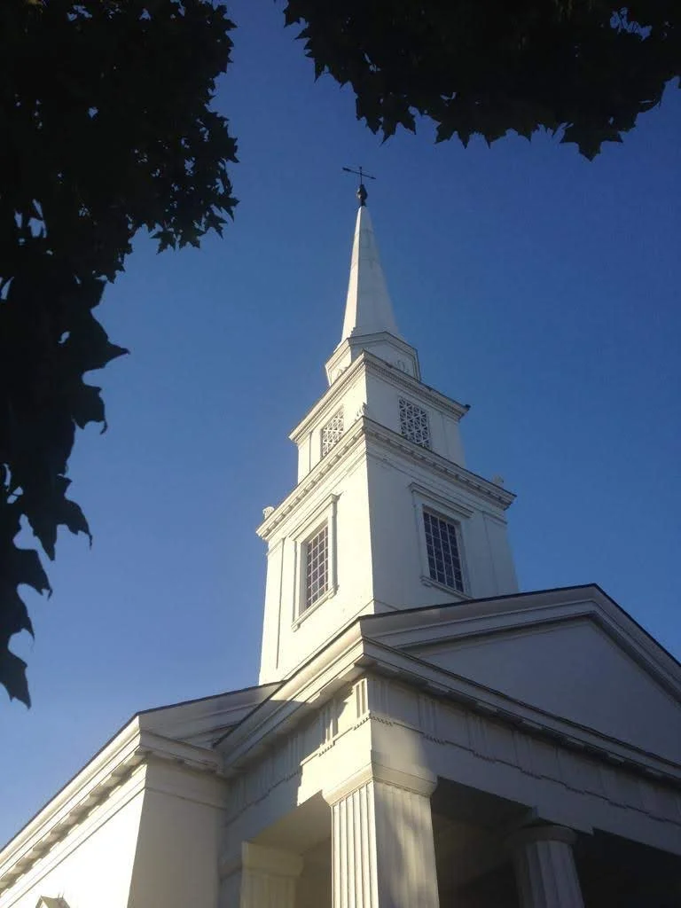 White church steeple with a cross on top against a clear blue sky, framed by tree branches.