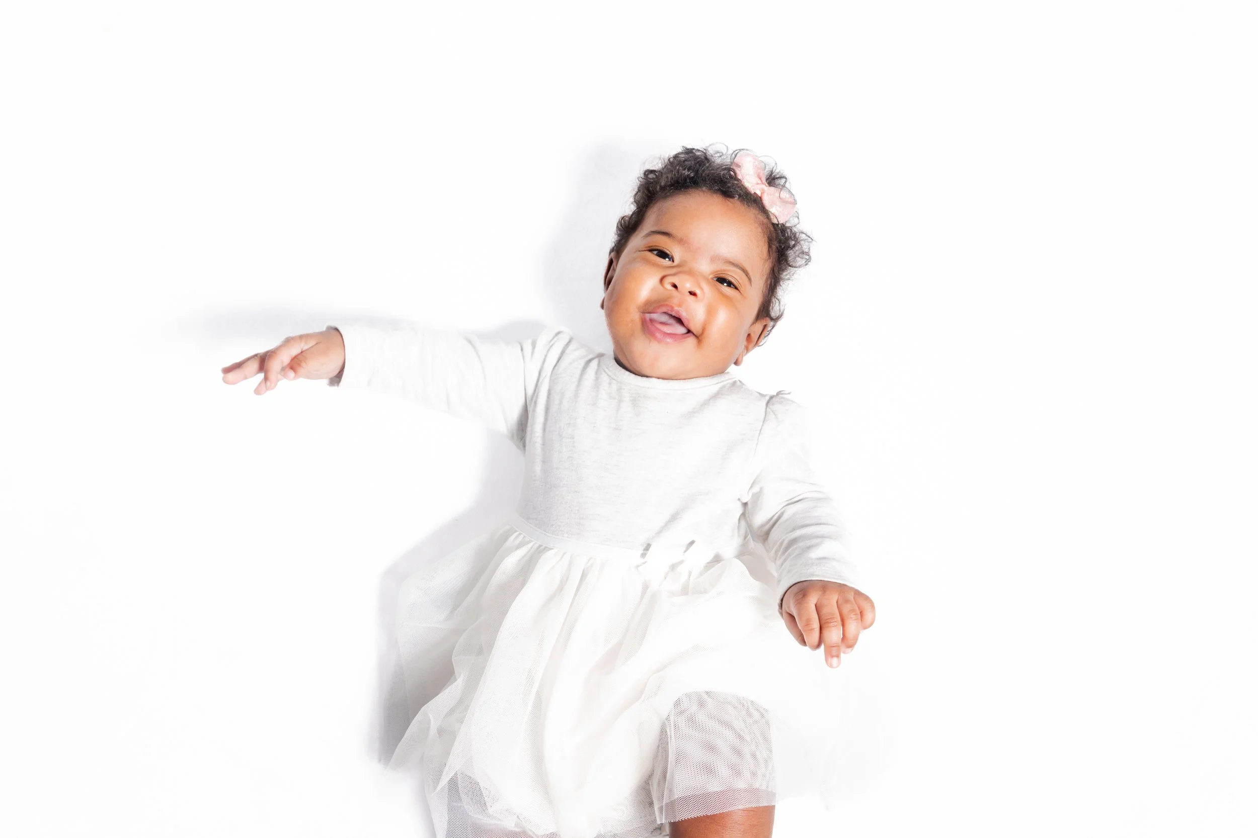 Happy toddler girl with curly hair wearing a white dress and pink bow, smiling and lying on a white background.