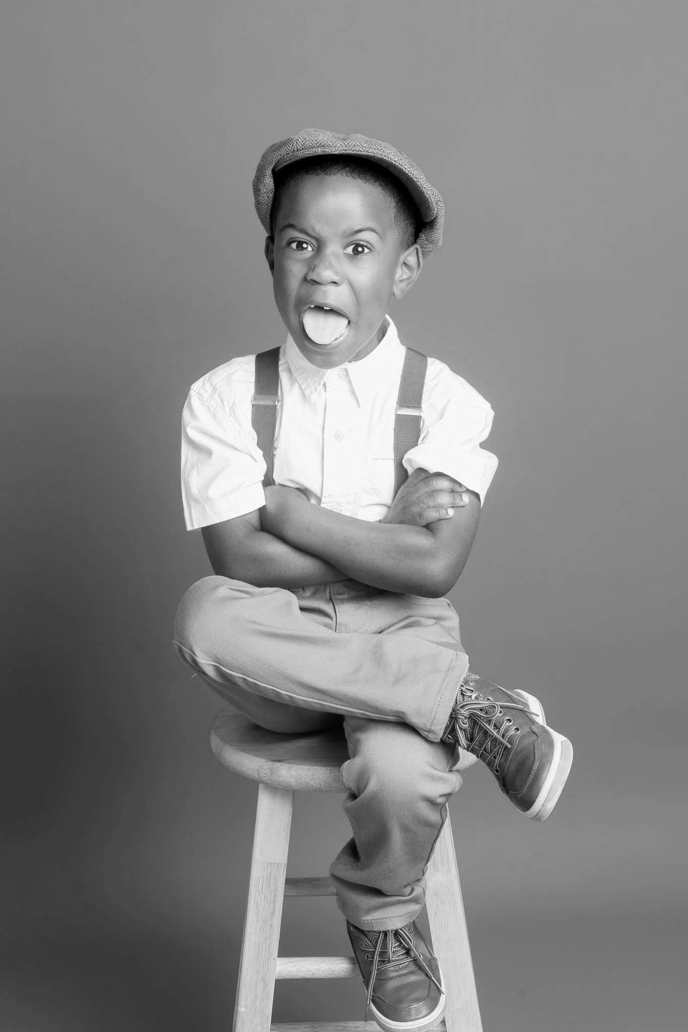 A young boy sitting on a stool with his arms crossed, wearing a hat, a button-up shirt, suspenders, pants, and boots, making a playful facial expression with tongue out.