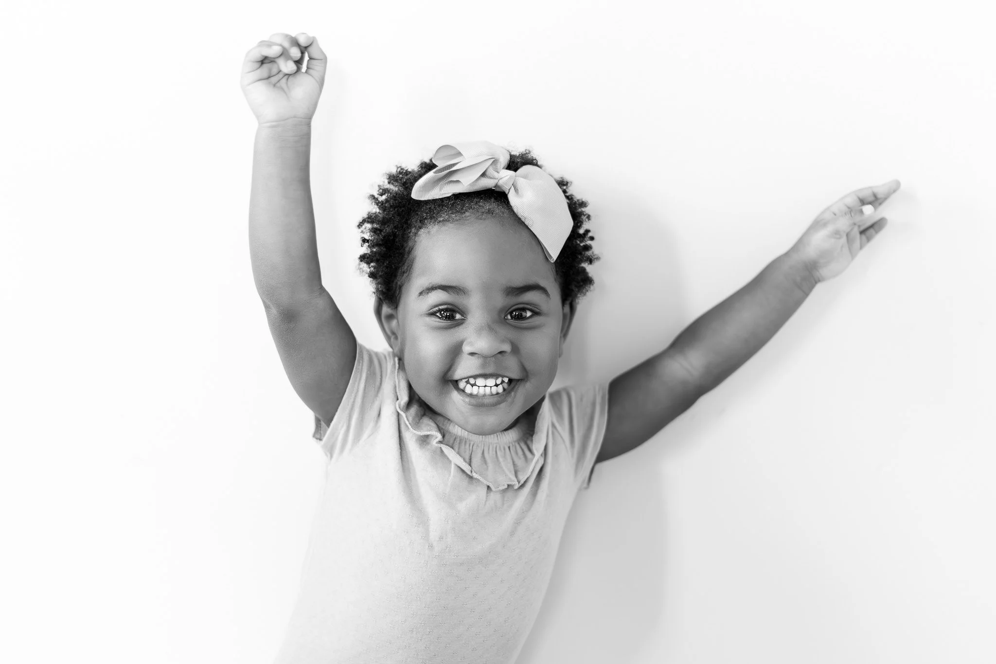 Black and white photo of a smiling young girl with curly hair, wearing a bow headband and a ruffled shirt, raising both arms joyfully.
