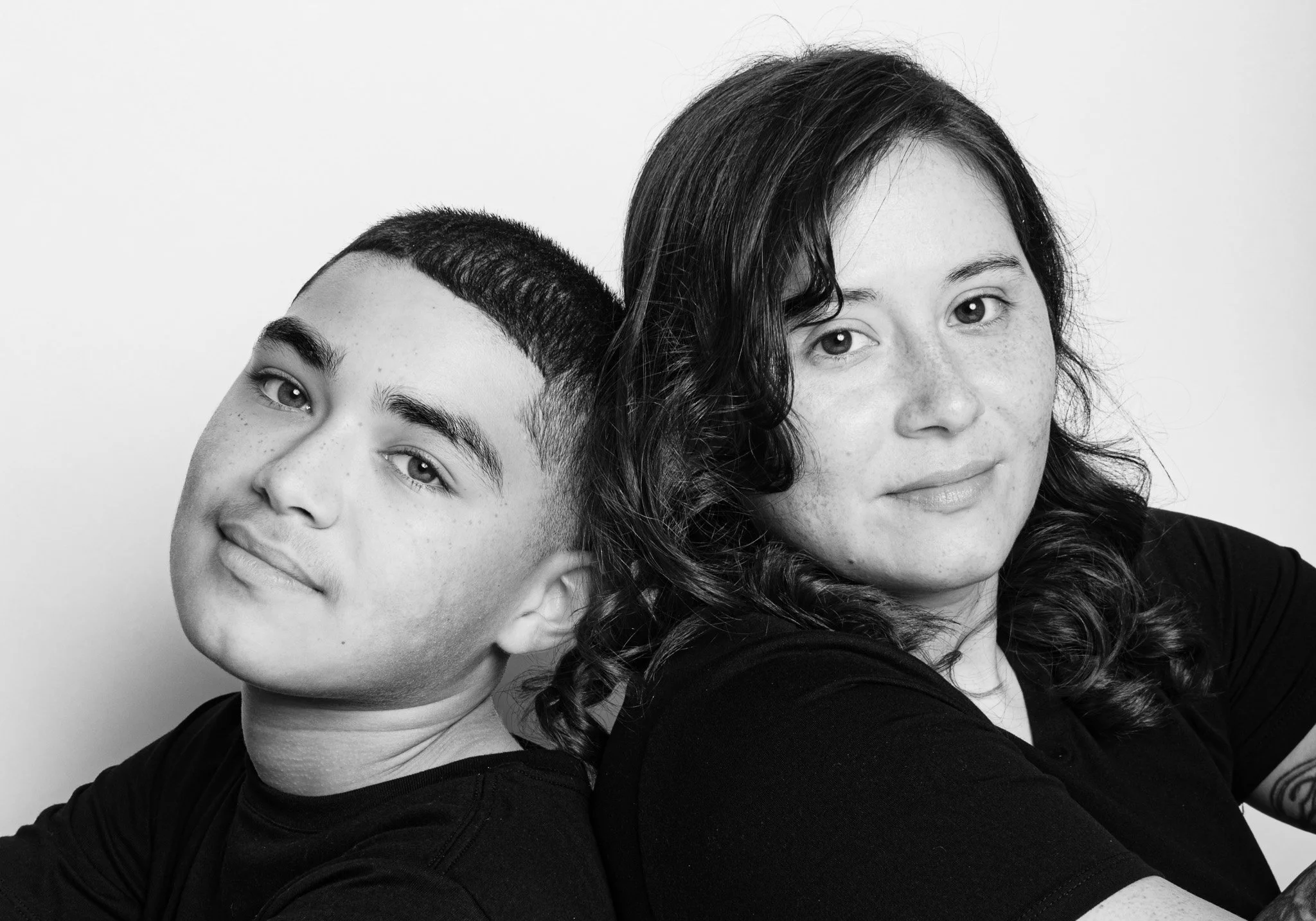 Black and white portrait of a teenage boy and a woman, sitting closely with the boy leaning his head back against the woman's shoulder, both smiling gently, against a plain background.
