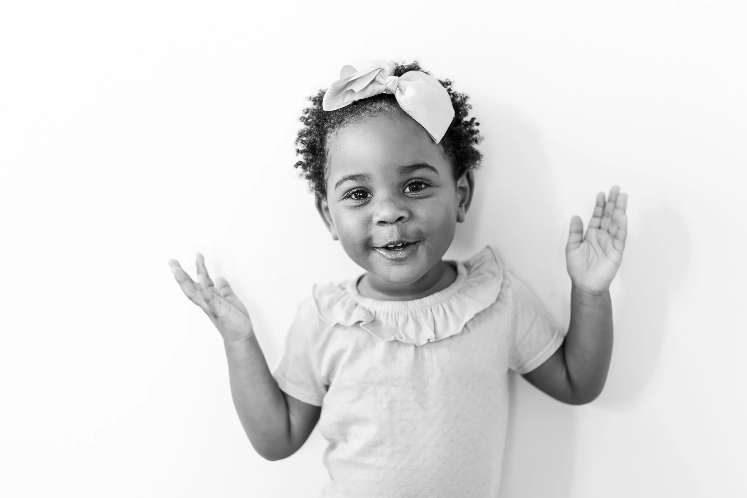 Black and white photo of a young girl with curly hair wearing a headband with a bow smiling and holding her hands up.