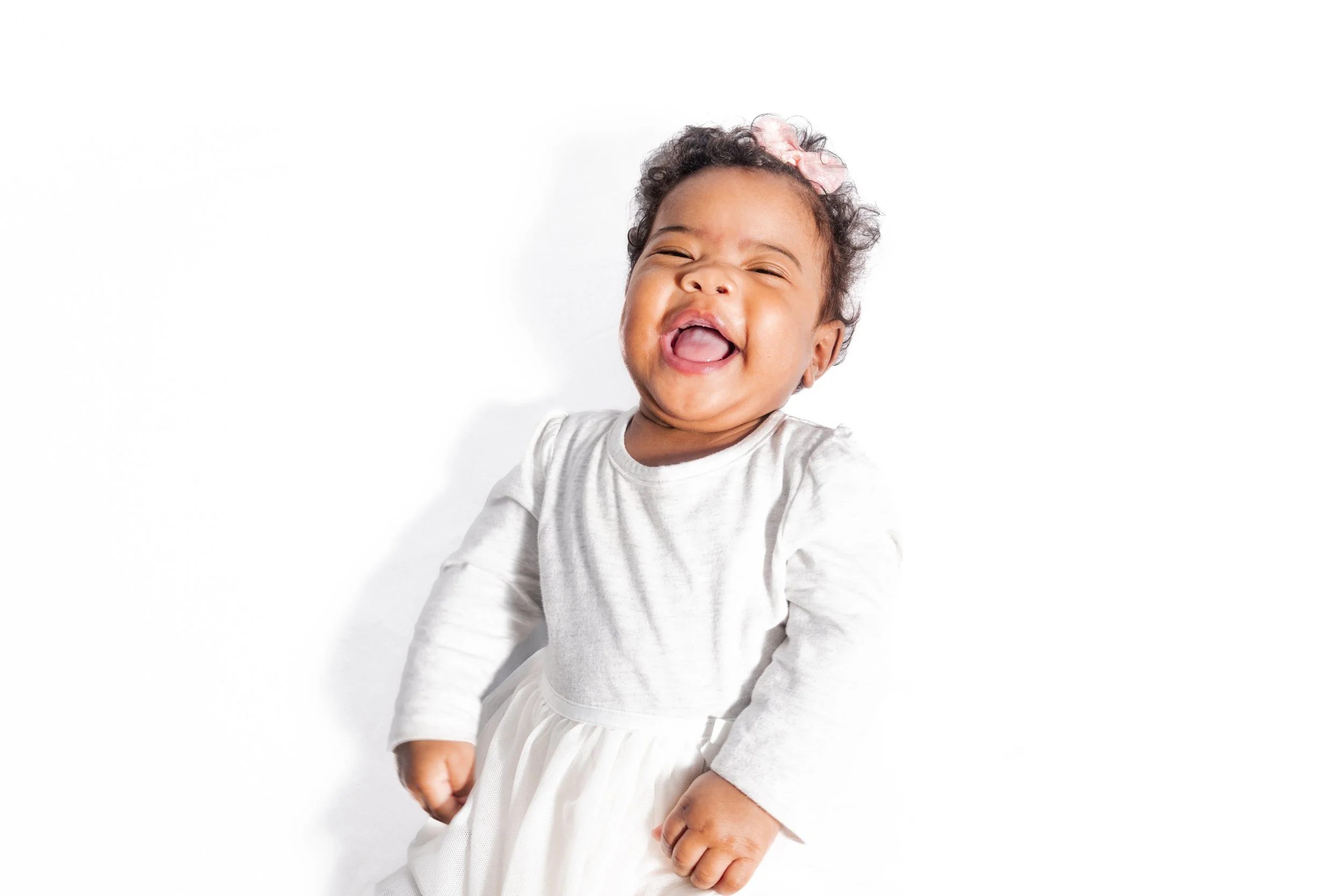 A happy toddler girl with curly hair and pink bow, smiling and laughing, wearing a white long-sleeve shirt and dress, standing against a white background.