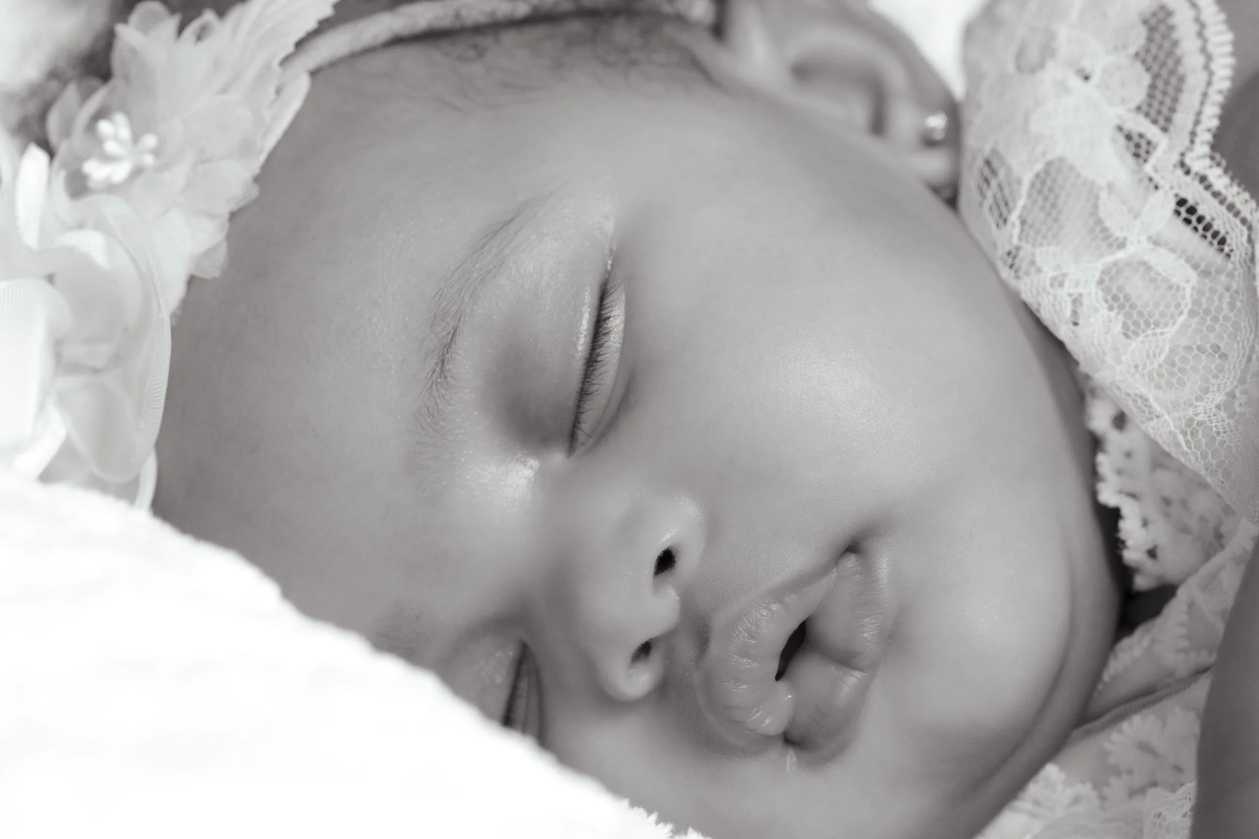 Close-up of a sleeping baby's face, in black and white, with delicate features and closed eyes, wearing a lace bonnet.