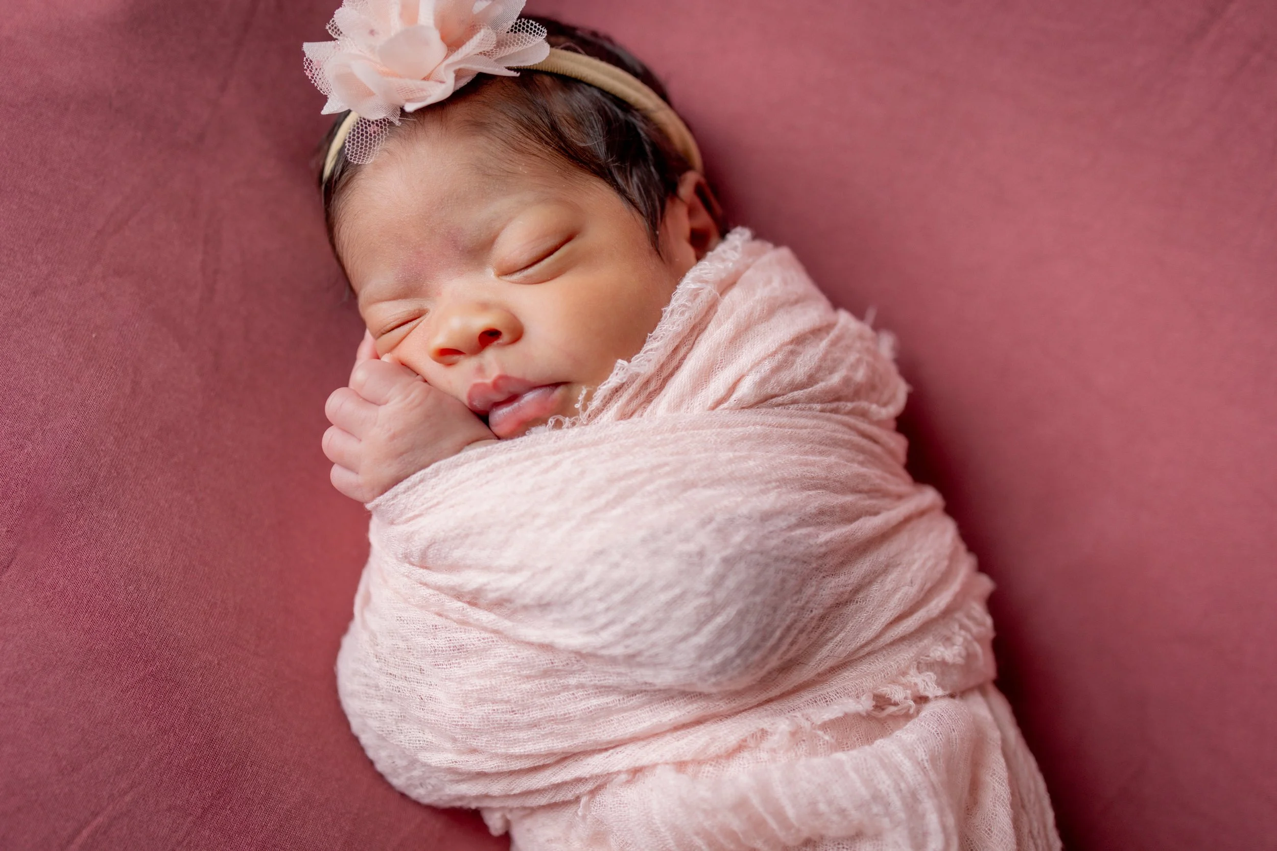 A newborn baby wrapped in a pink blanket, sleeping on a pink surface, wearing a headband with a large pink flower.