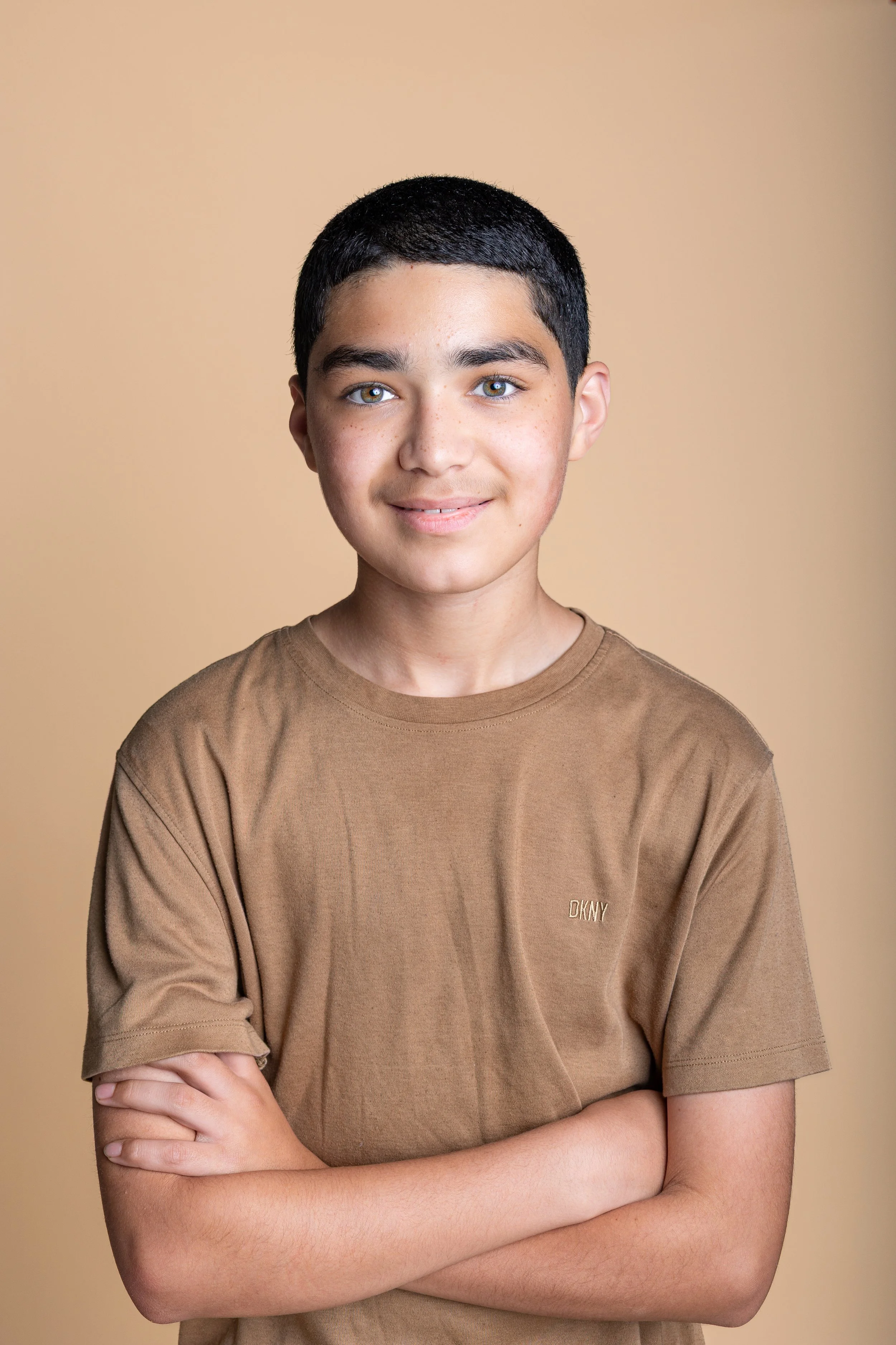 A young boy with short black hair and blue eyes, wearing a brown DKNY t-shirt, standing with arms crossed against a beige background.