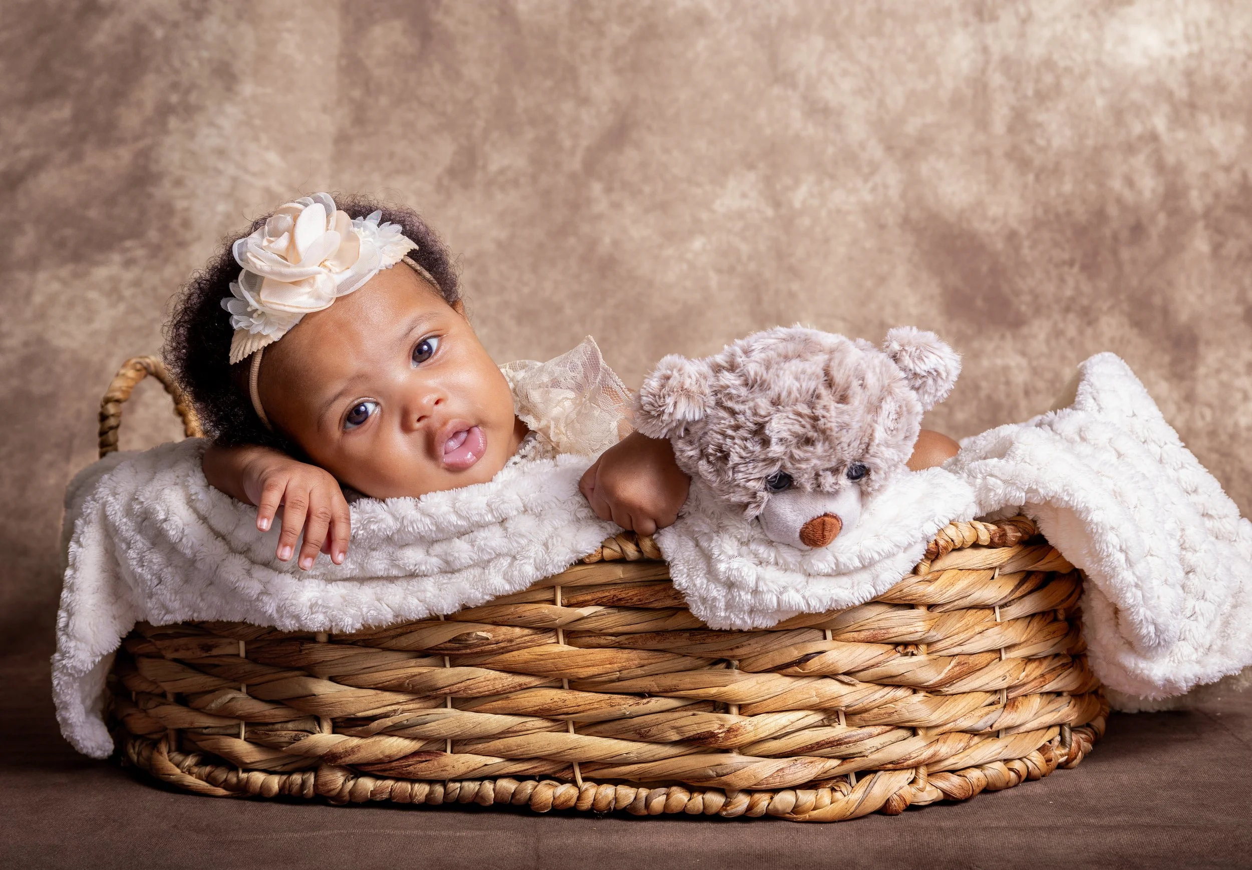 A young girl with a floral headband lying in a wicker basket with a teddy bear, wrapped in a plush white blanket, against a neutral background.