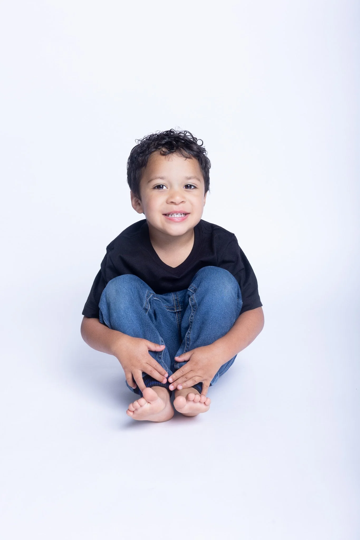 A young boy with curly dark hair, wearing a black t-shirt and blue jeans, sitting barefoot on a white surface and smiling at the camera.