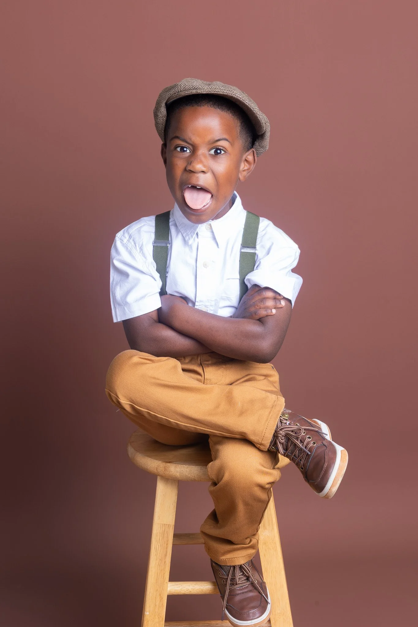 A young boy sitting on a wooden stool with arms crossed, wearing a white shirt, brown pants, boots, and a hat, making a surprised or excited facial expression with mouth open and tongue out, against a plain brown background.
