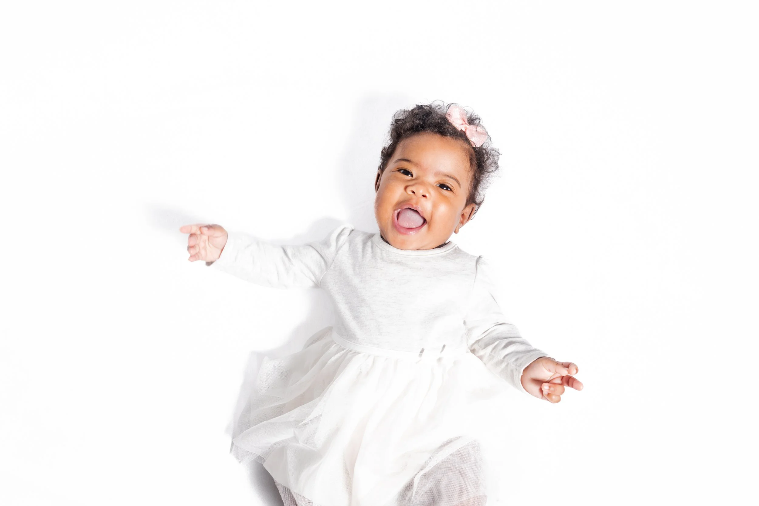 Smiling baby girl with a pink bow in her curly hair, wearing a white dress, lying on a white background, with arms outstretched