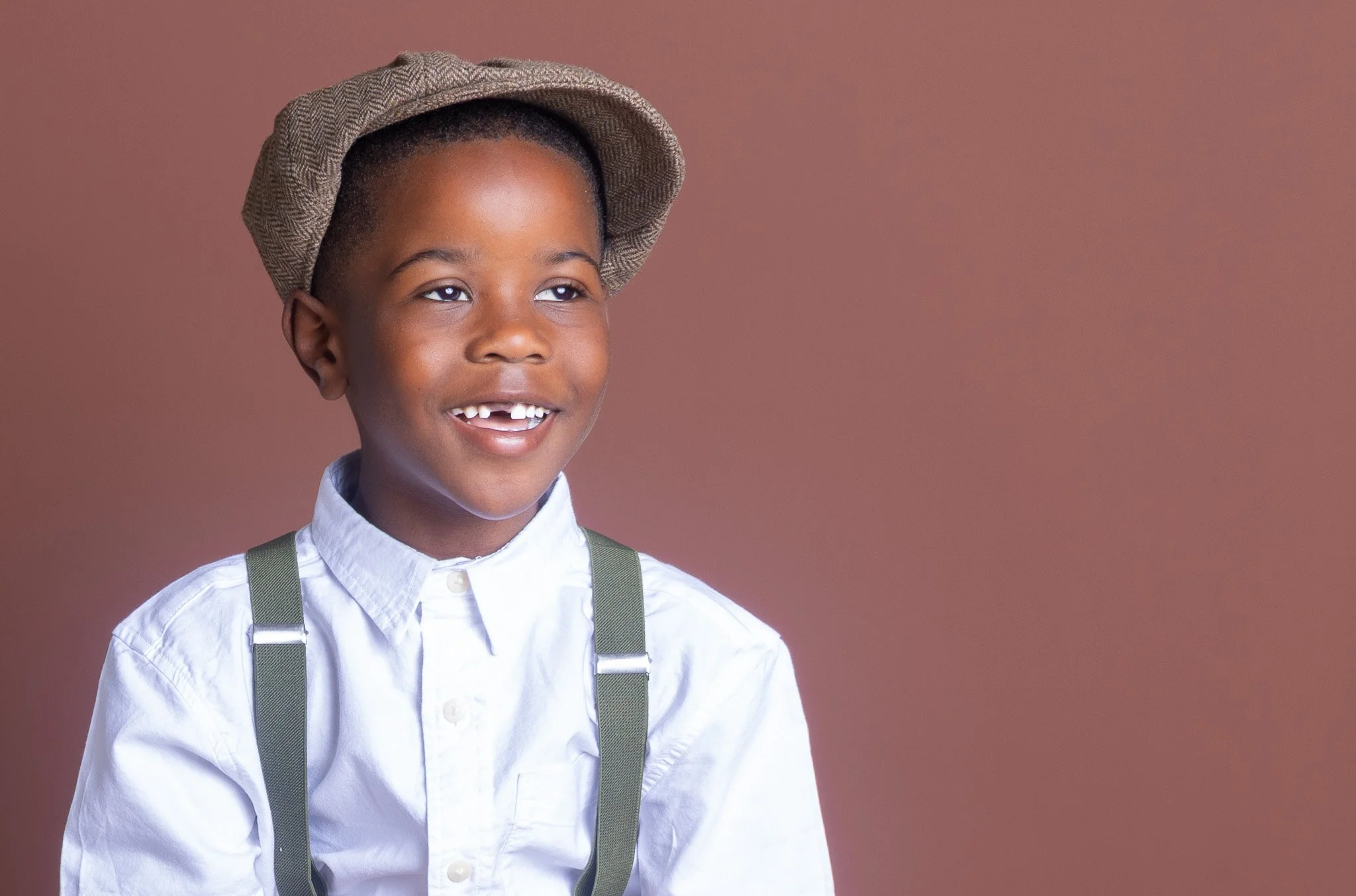 Young boy wearing a white shirt, suspenders, newsboy cap, smiling, against a plain brown background.