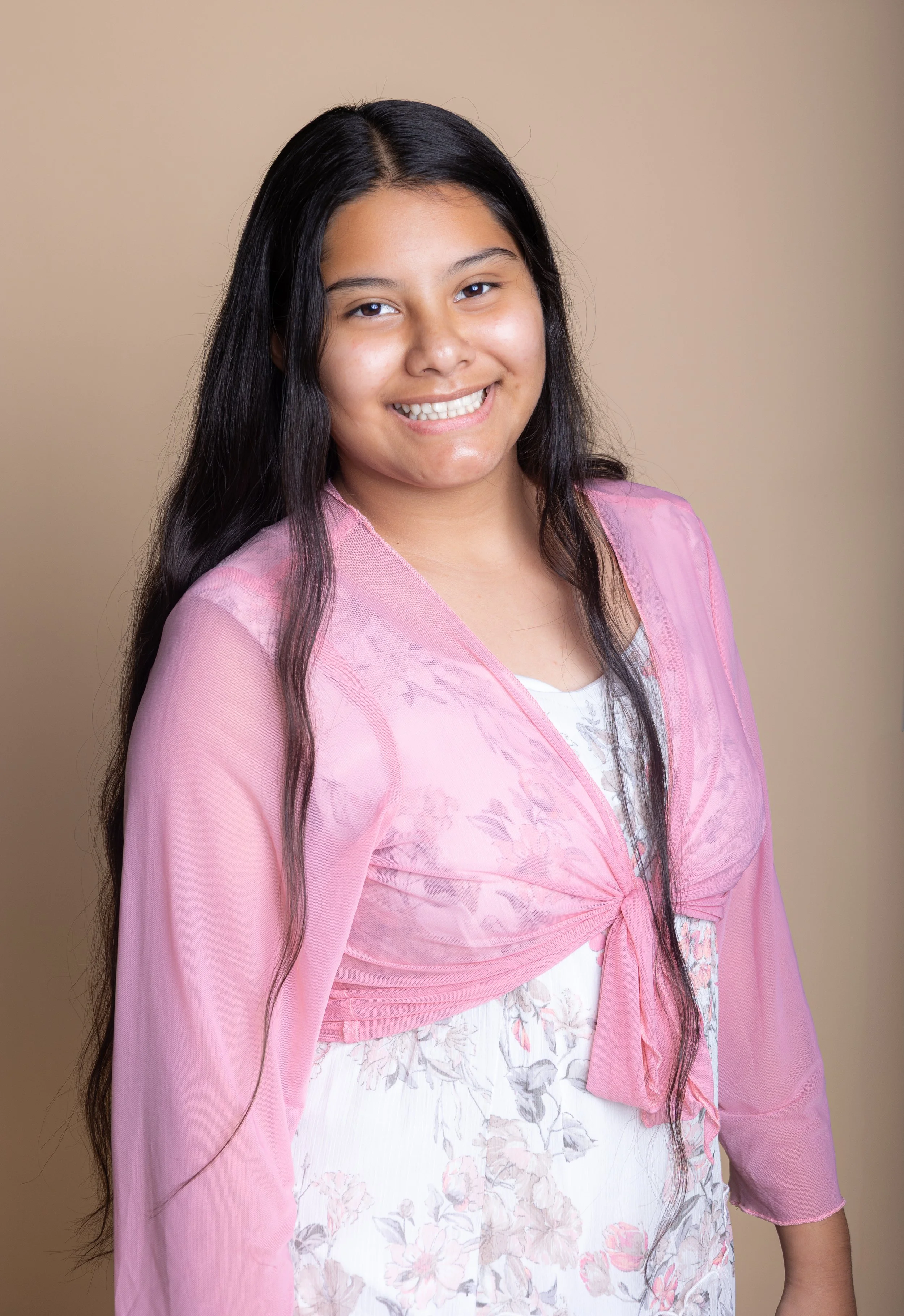 Young woman with long dark hair smiling, wearing a pink sheer jacket over a white floral dress, standing against a beige background.