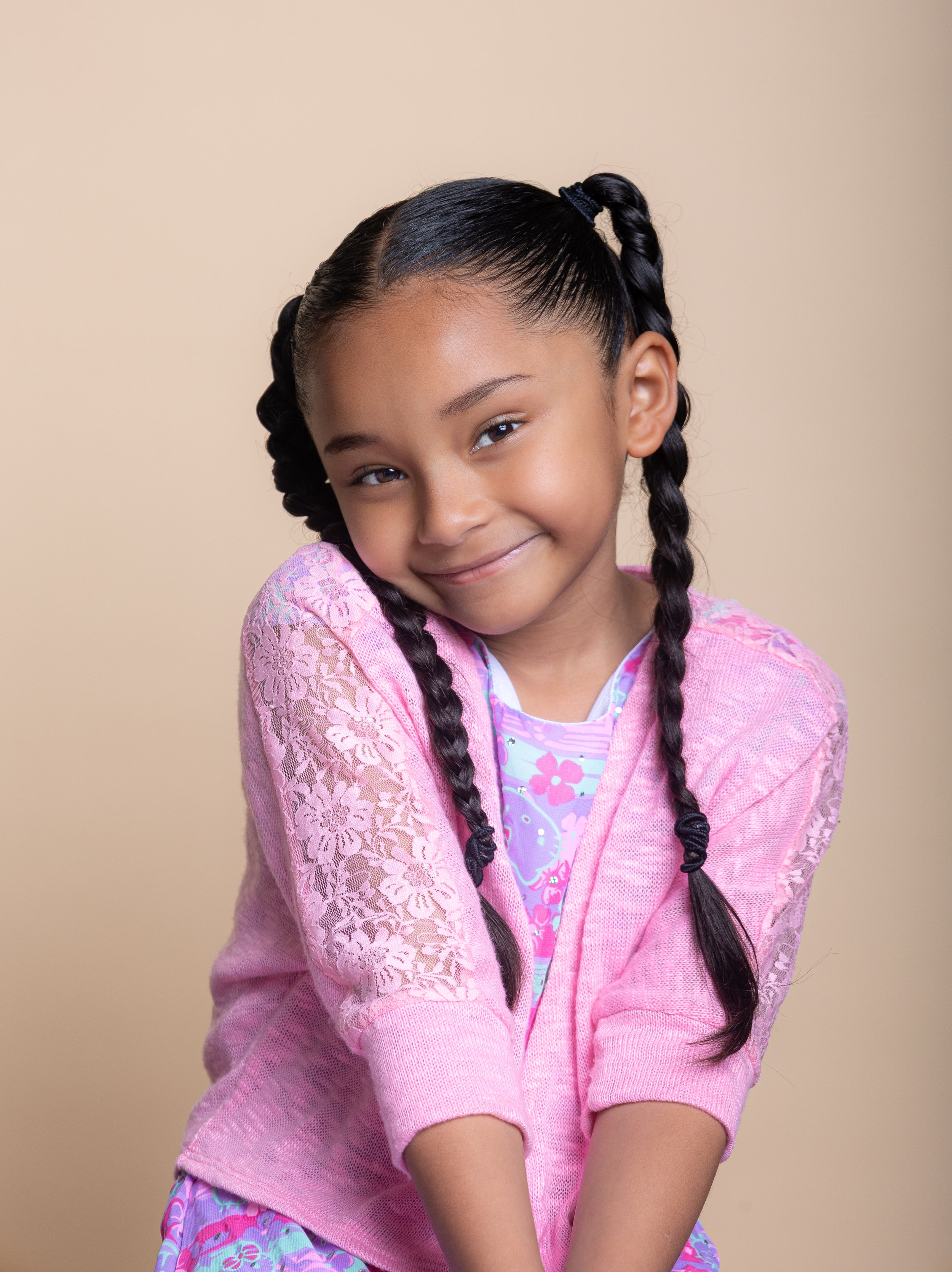 A young girl with braided hair smiling and tilting her head to the side, wearing a pink lace hoodie and a colorful dress against a beige background.
