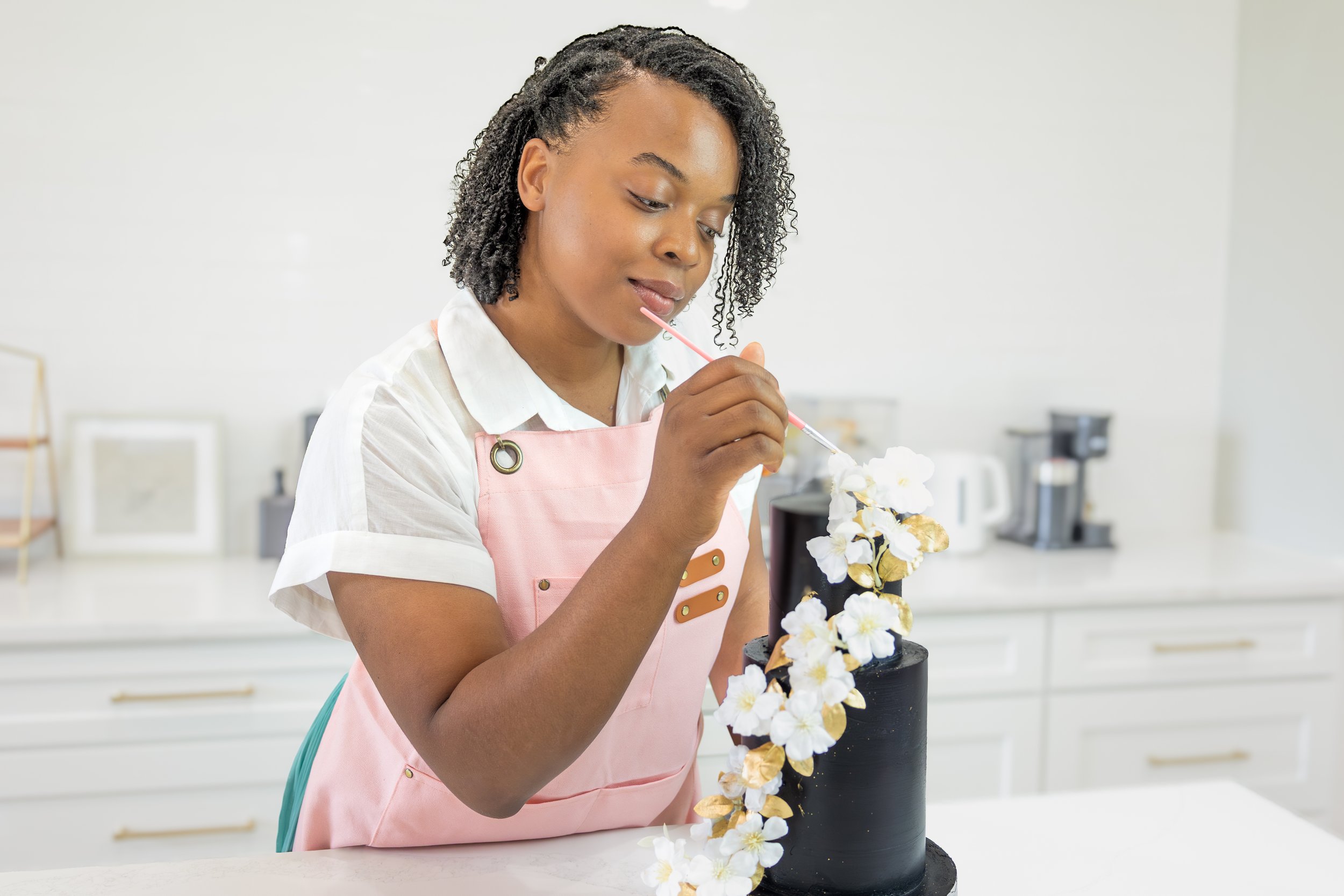 A woman decorating a black cake with white and gold floral decorations in a kitchen.