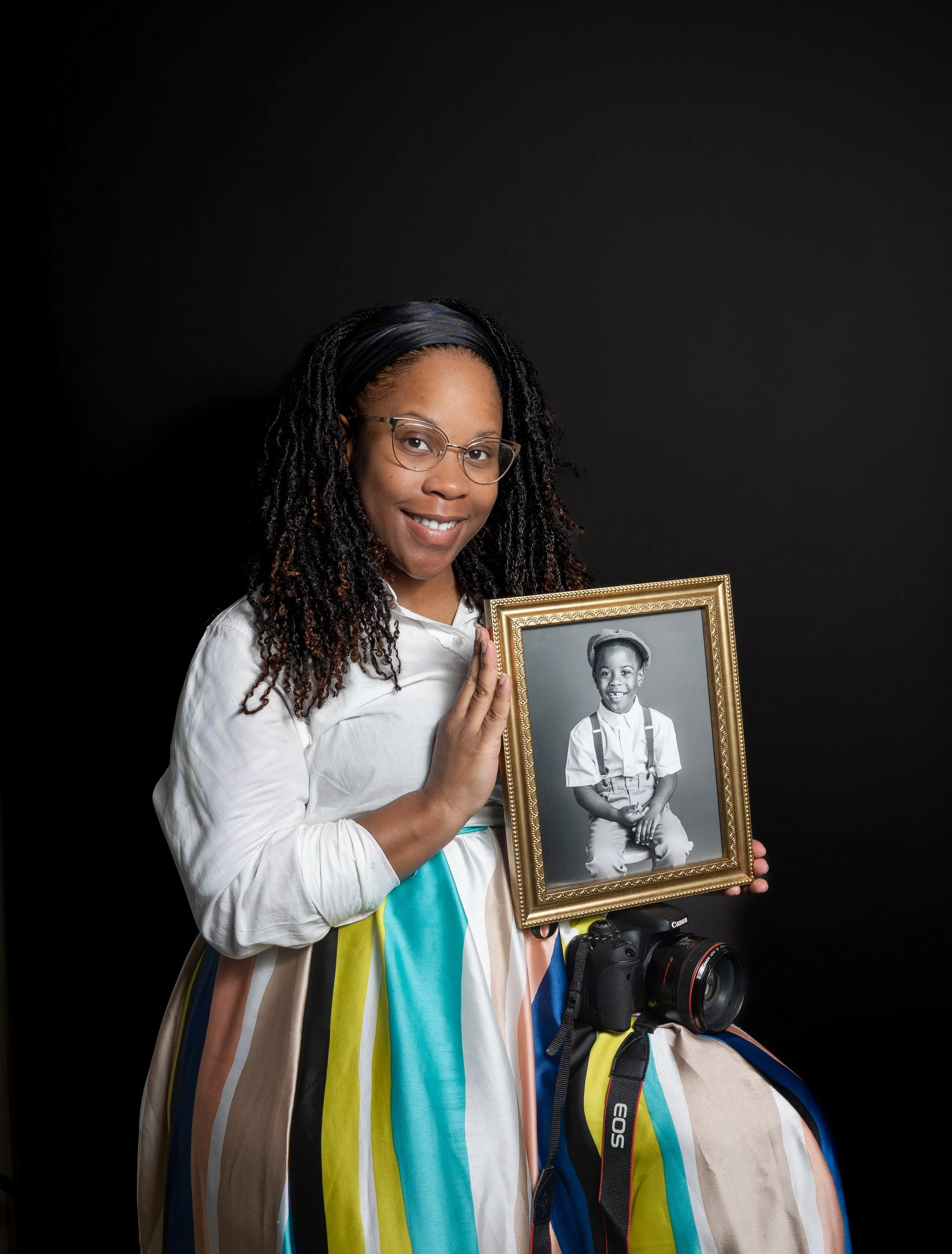 A woman with glasses and long curly hair, wearing a white blouse and a colorful striped skirt, is smiling and holding a framed black-and-white childhood photograph of herself.