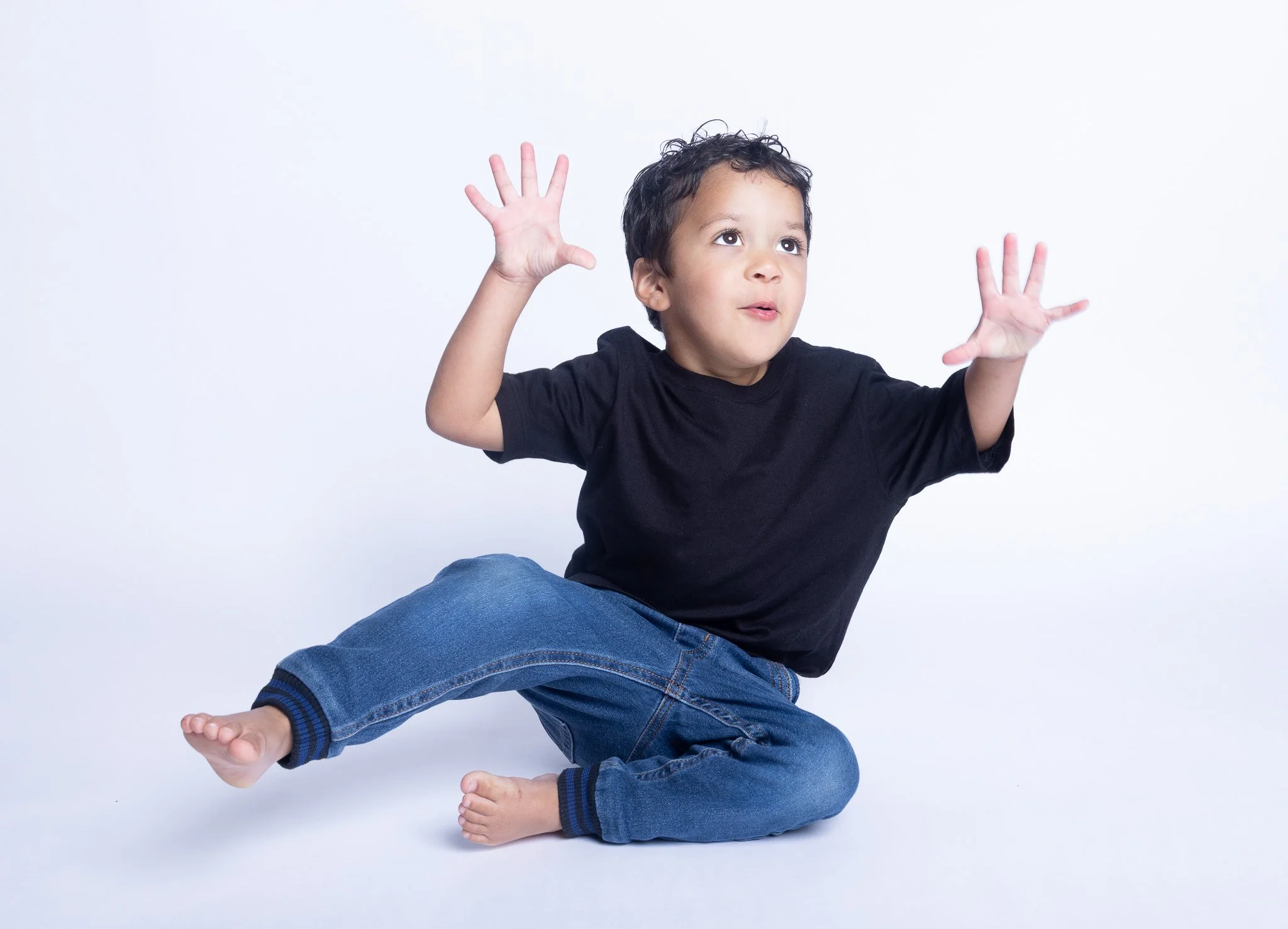 Young boy sitting on the floor with one leg crossed over the other, wearing a black T-shirt and jeans, with hands raised and a curious expression.