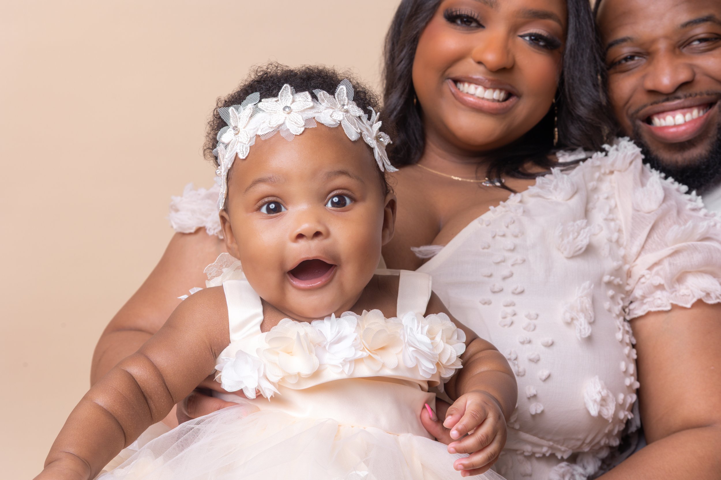 Smiling African-American family with a mother, father, and a toddler girl dressed in light-colored, floral-embellished dresses, sitting together against a neutral background.
