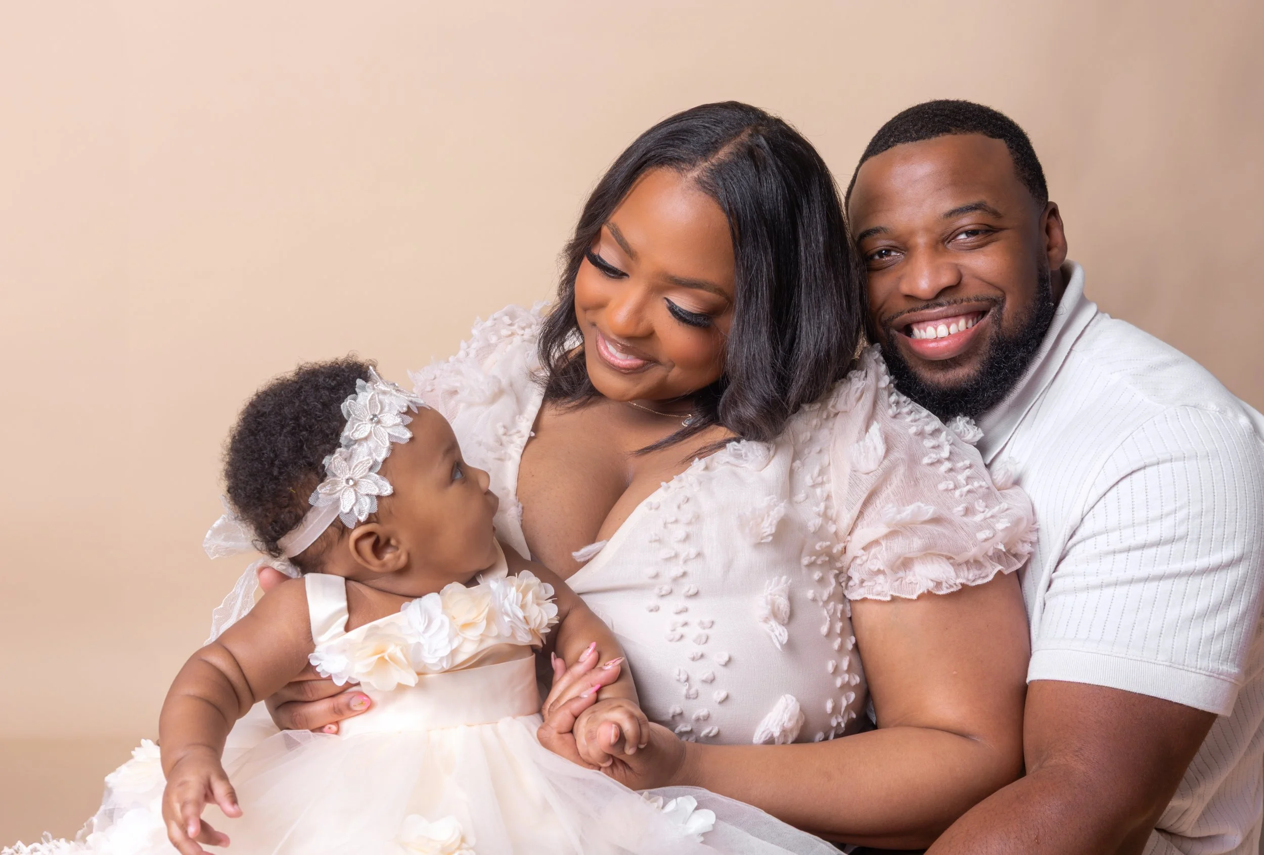 Happy family of three celebrating and smiling, with the mother holding a baby girl in a white dress and floral headband, and the father embracing them, set against a neutral background.