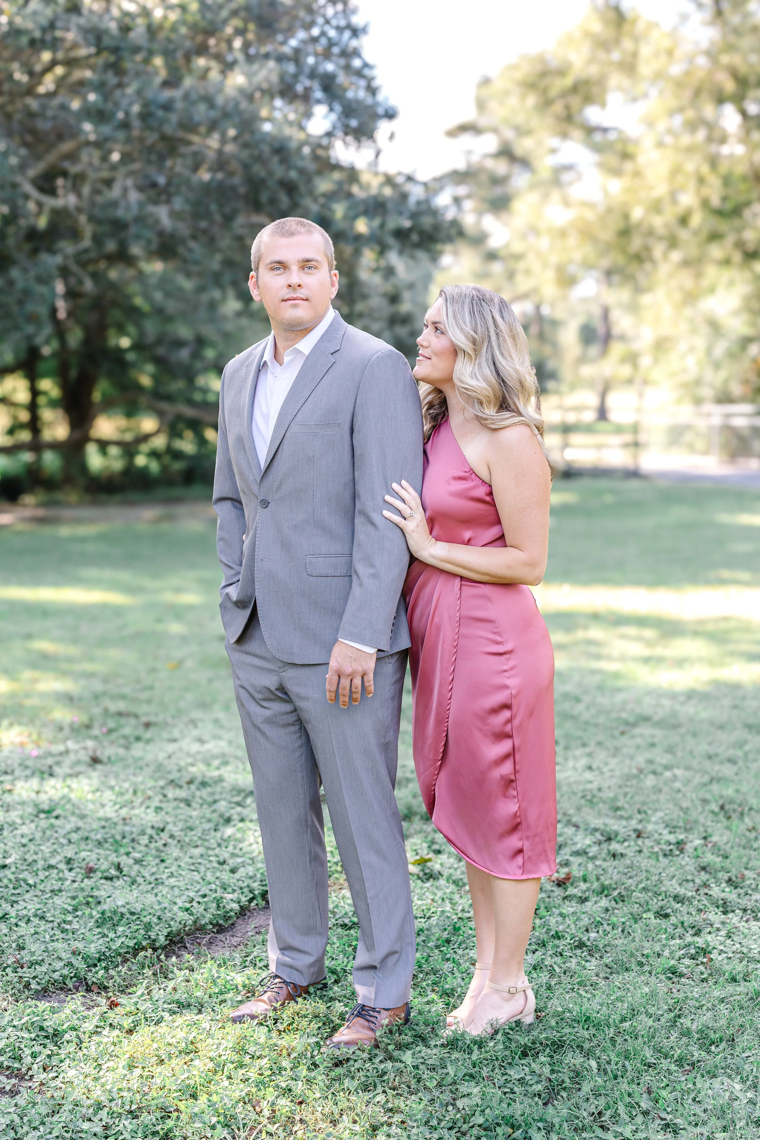 A man in a gray suit standing outdoors with a woman in a pink dress leaning on his shoulder and looking at him. They are on a grassy area with trees in the background.