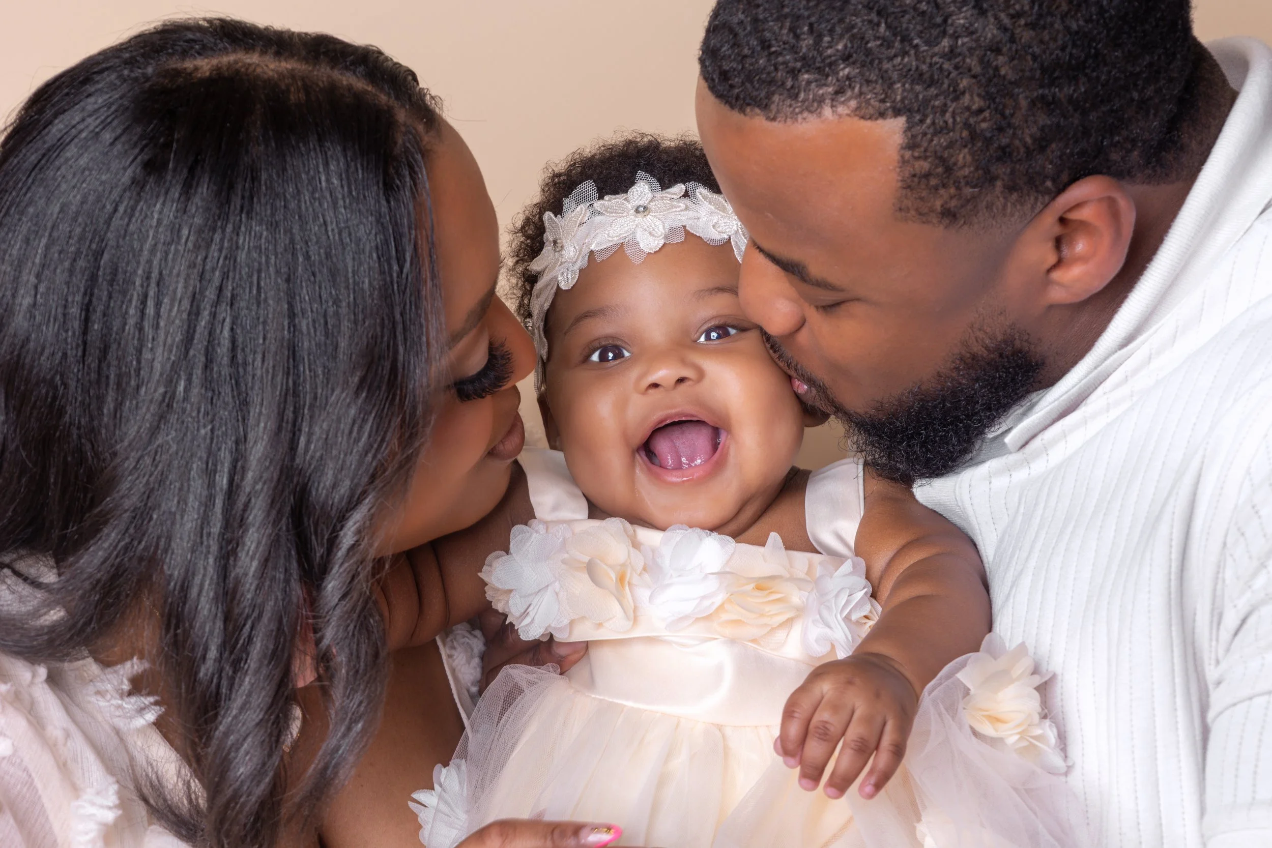A happy African-American family with a woman, man, and baby girl, sharing a joyful moment as they kiss the baby's cheeks.