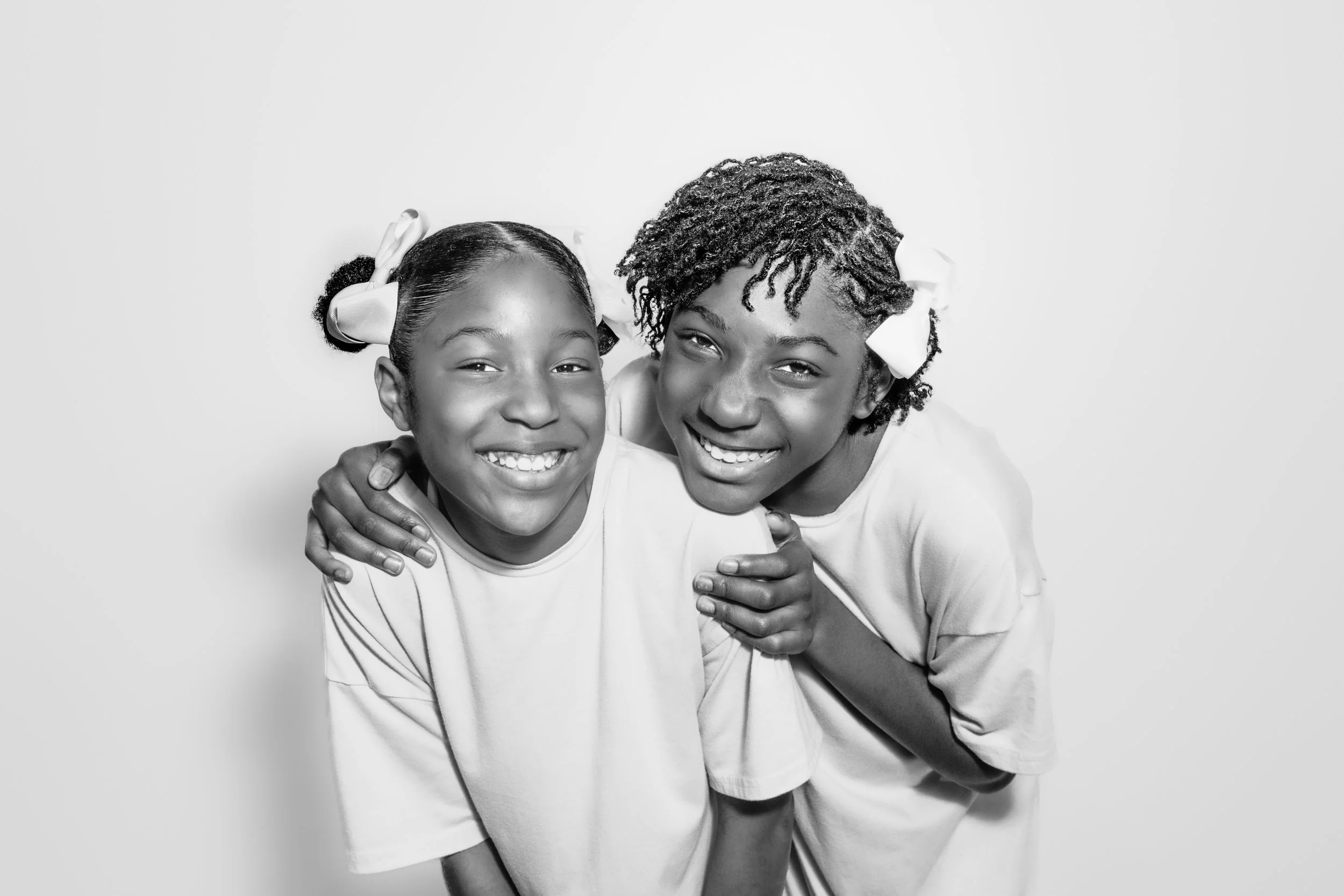 Two young girls smiling and hugging each other in front of a plain background, dressed in light-colored shirts, with hair accessories.