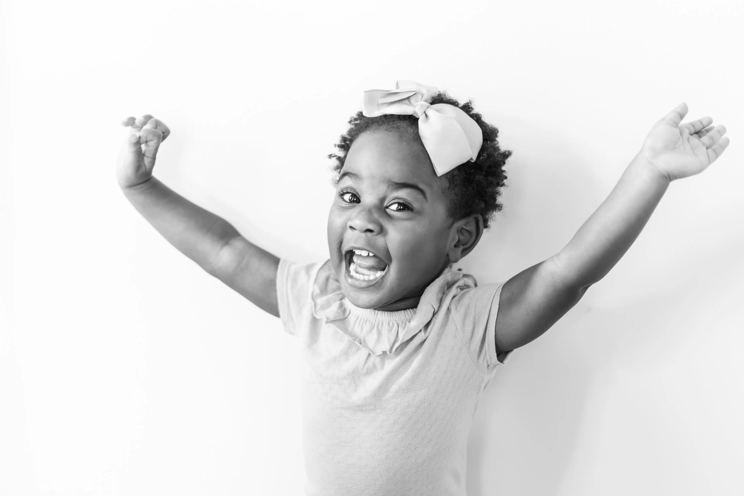 A young girl with curly hair, wearing a bow headband and a light-colored top, is smiling with her mouth open and raising her arms in a celebratory pose against a plain white background.