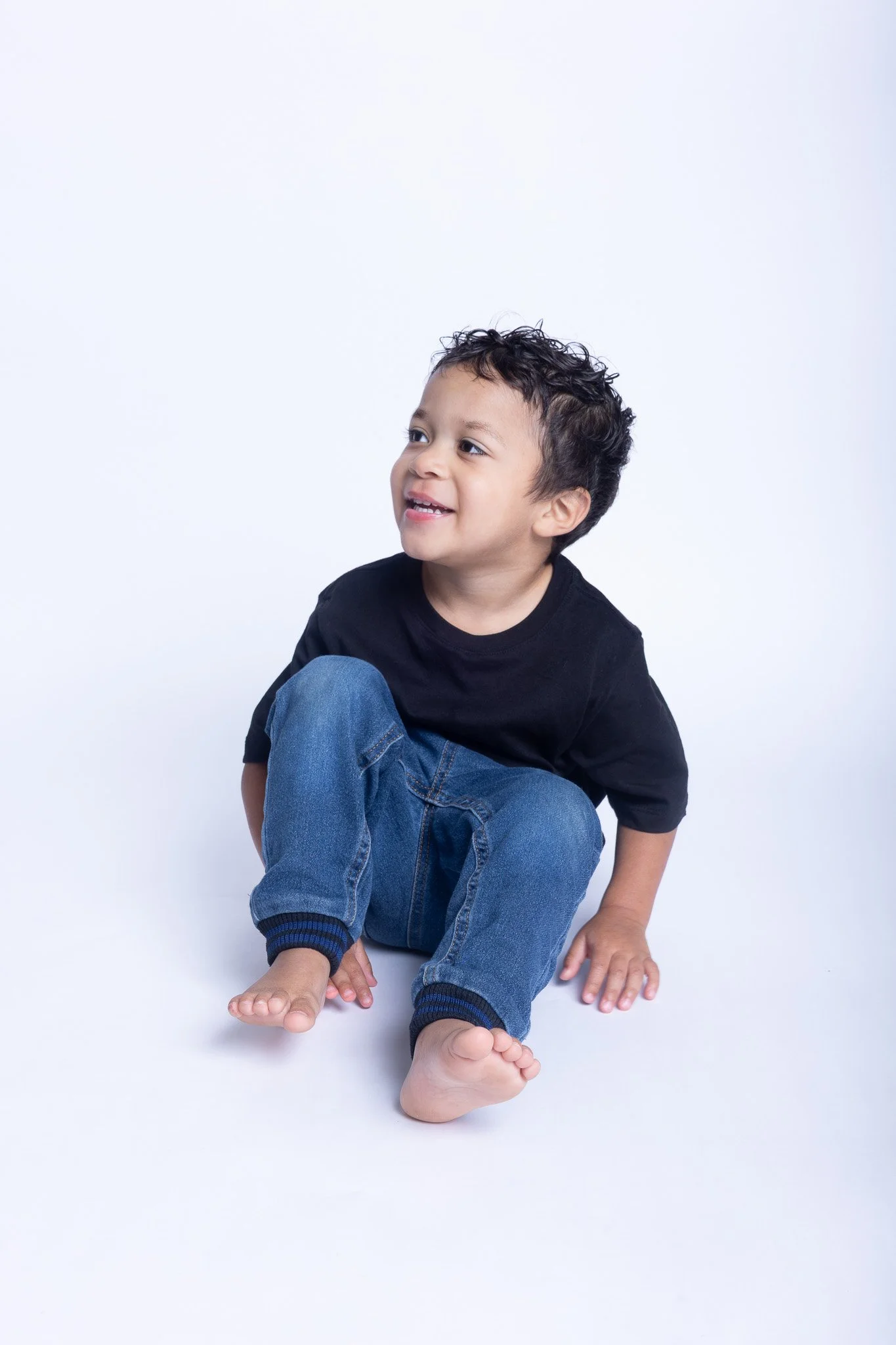 Young boy sitting on the floor, smiling, wearing a black T-shirt and blue jeans, with a white background.