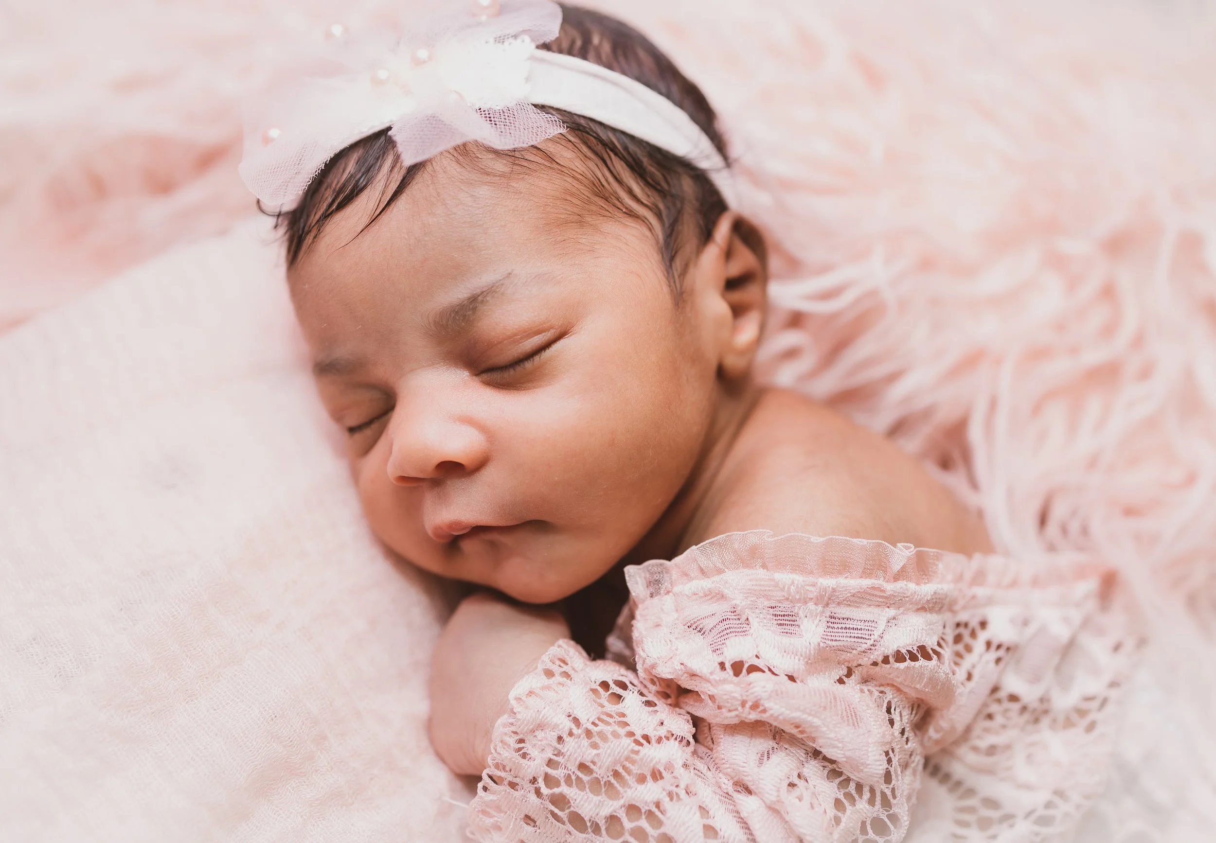 Close-up of a sleeping baby girl with a pink lace dress and a white headband with a pink bow, lying on a soft pink fluffy blanket.