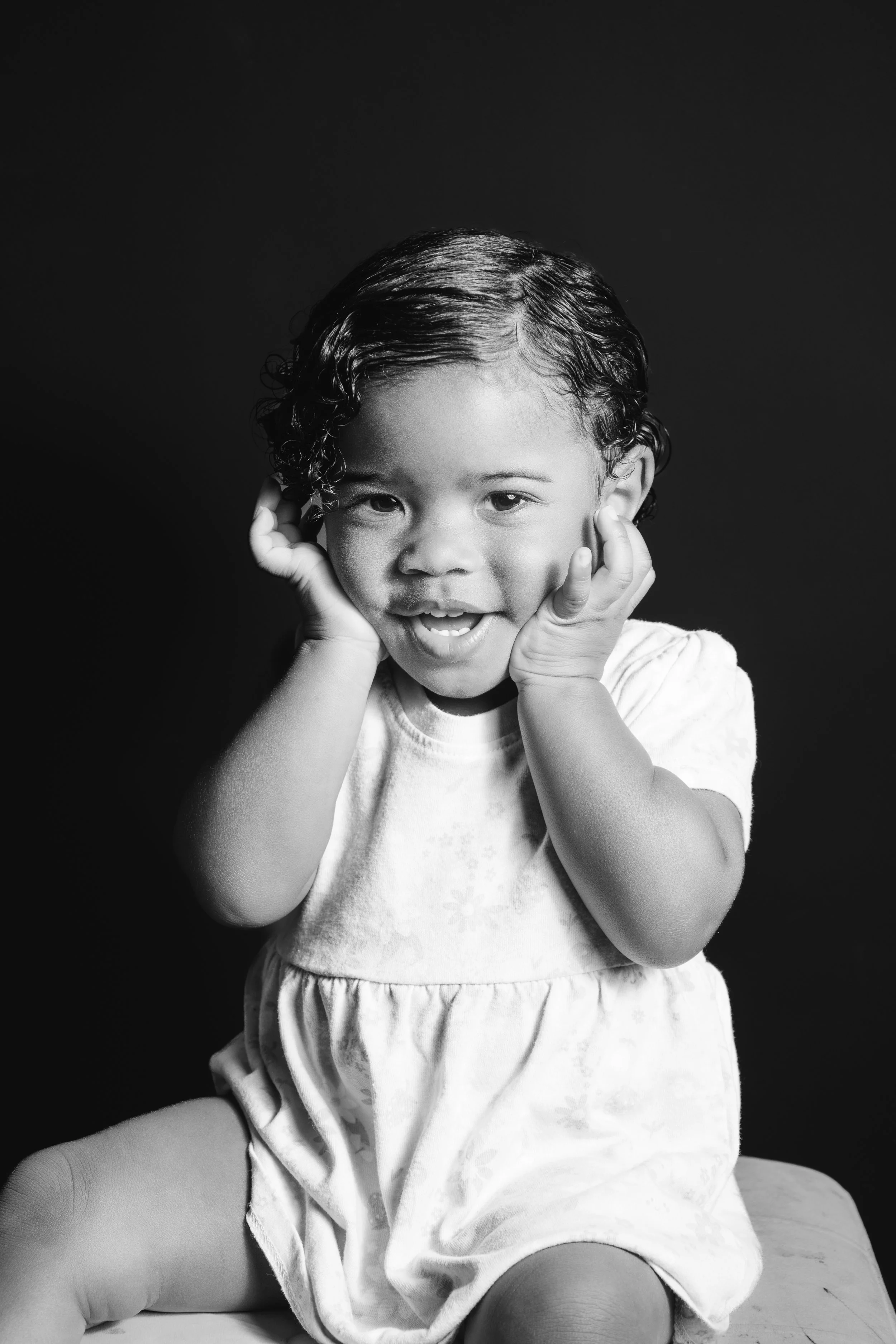 A young girl with curly hair smiling and holding her face with her hands, sitting against a dark background.