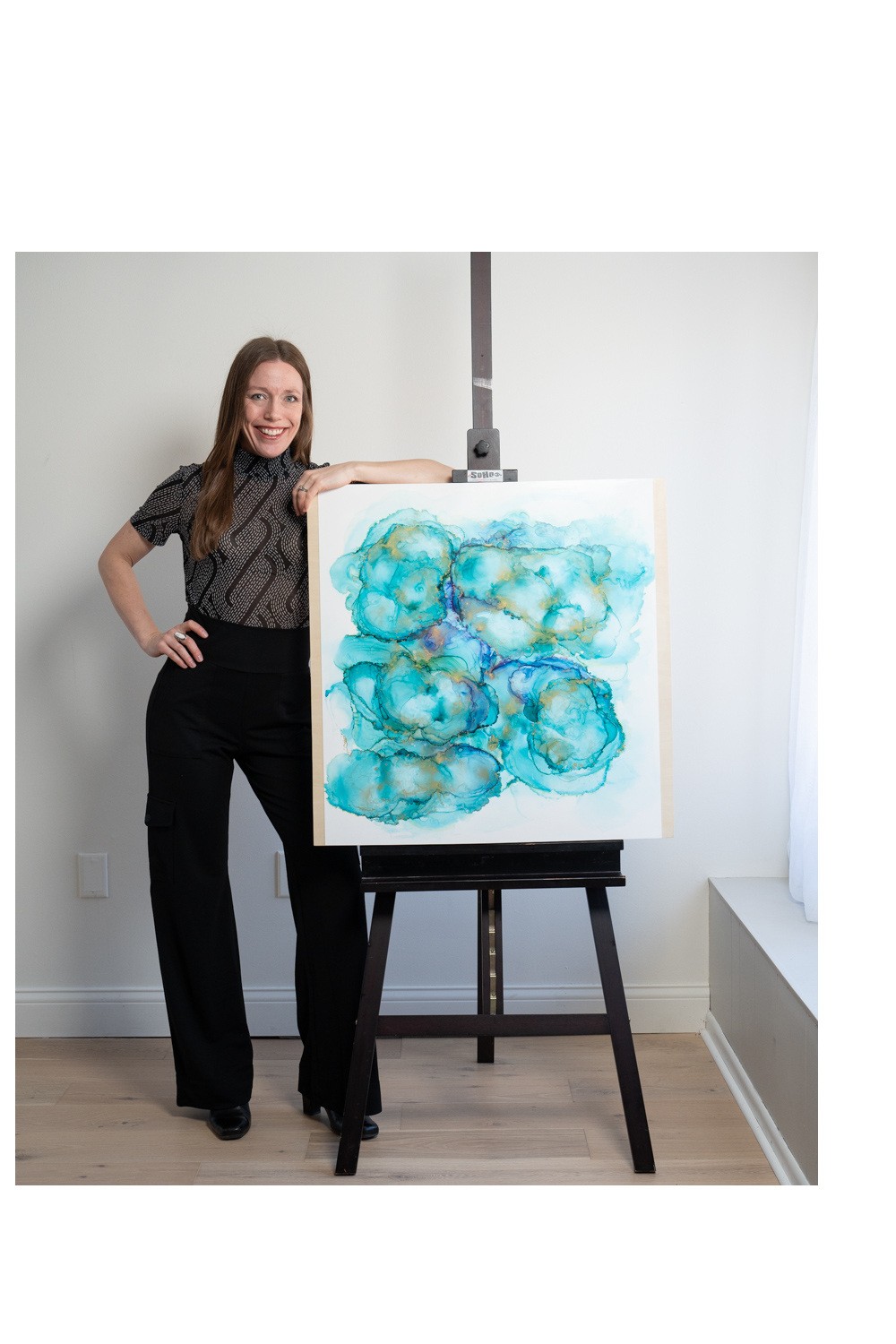 A woman standing next to an easel displaying an abstract watercolor painting with blue, teal, and gold colors.
