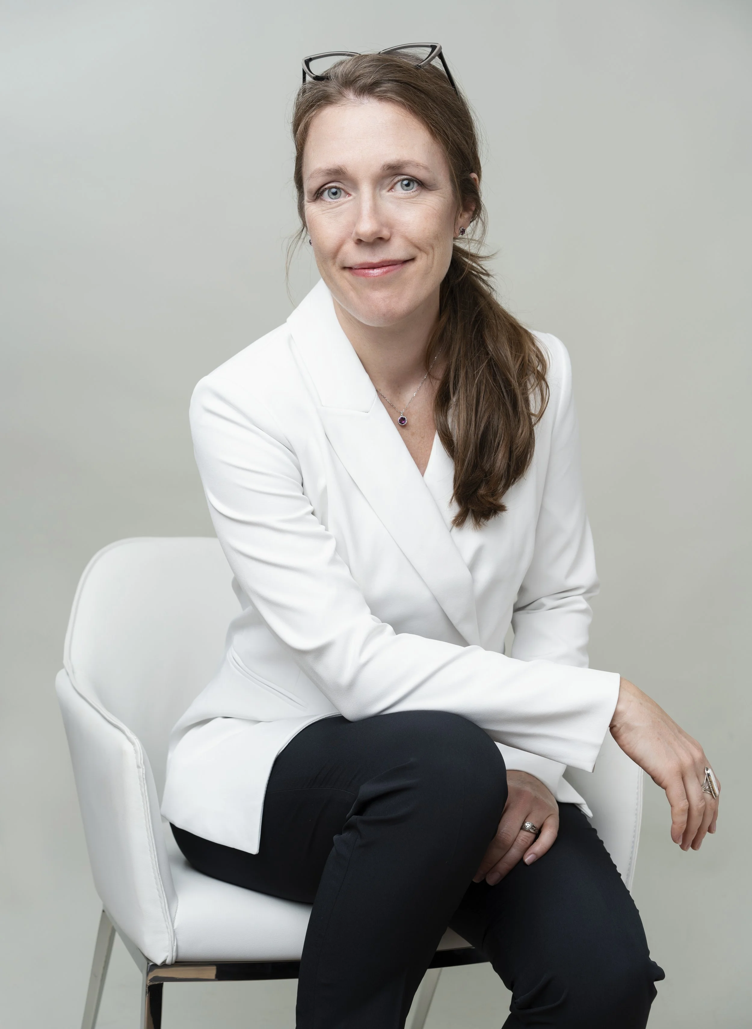 A woman with long brown hair and glasses on her head, wearing a white blazer, black pants, and jewelry, sitting on a white chair against a plain light-colored background.