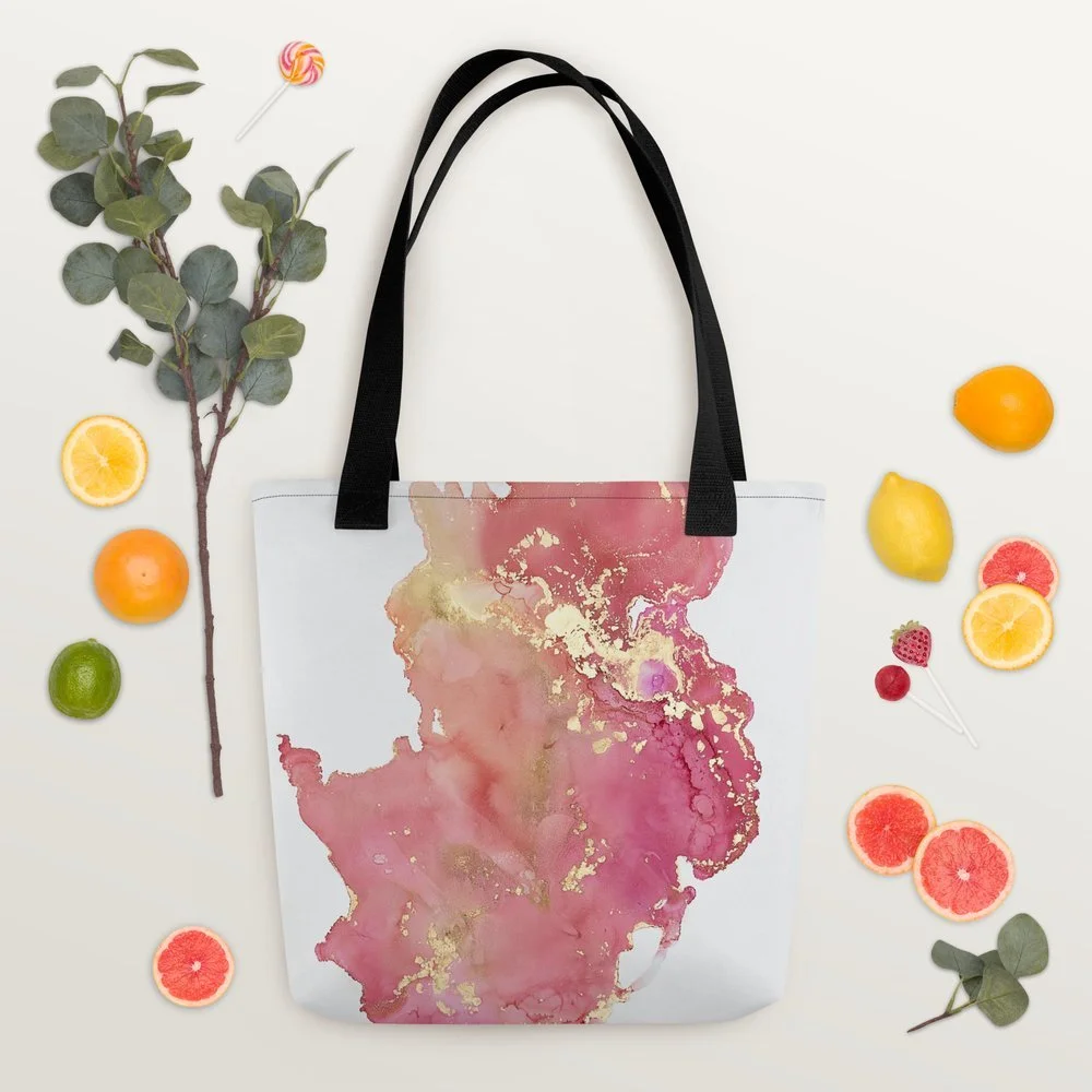 A white tote bag with pink and gold watercolor design, black handles, surrounded by various fruits including lemon, lime, orange, grapefruit slices, strawberries, cherries, and a branch with green leaves, arranged on a white background with a pink and yellow lollipop.
