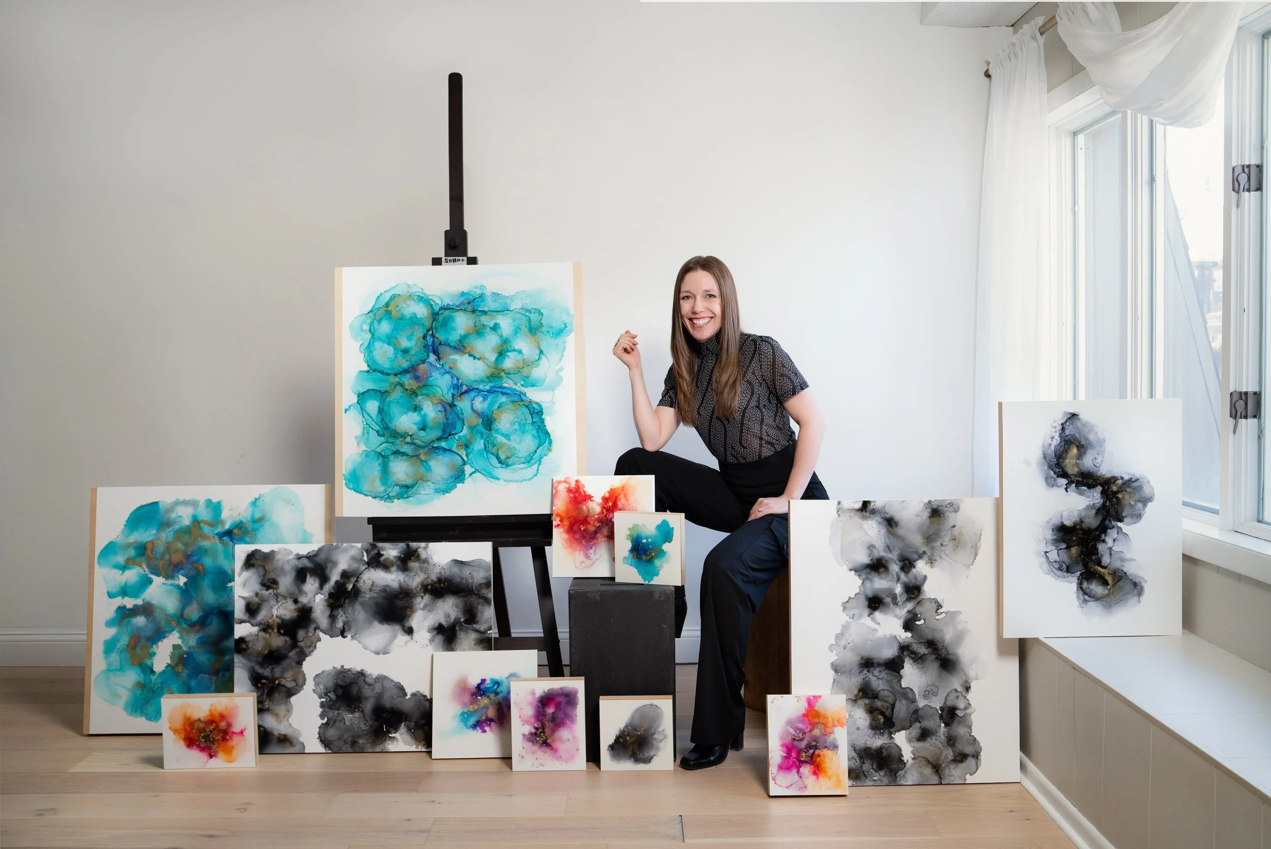 Artist sitting in art studio surrounded by abstract watercolor paintings in various colors and sizes.