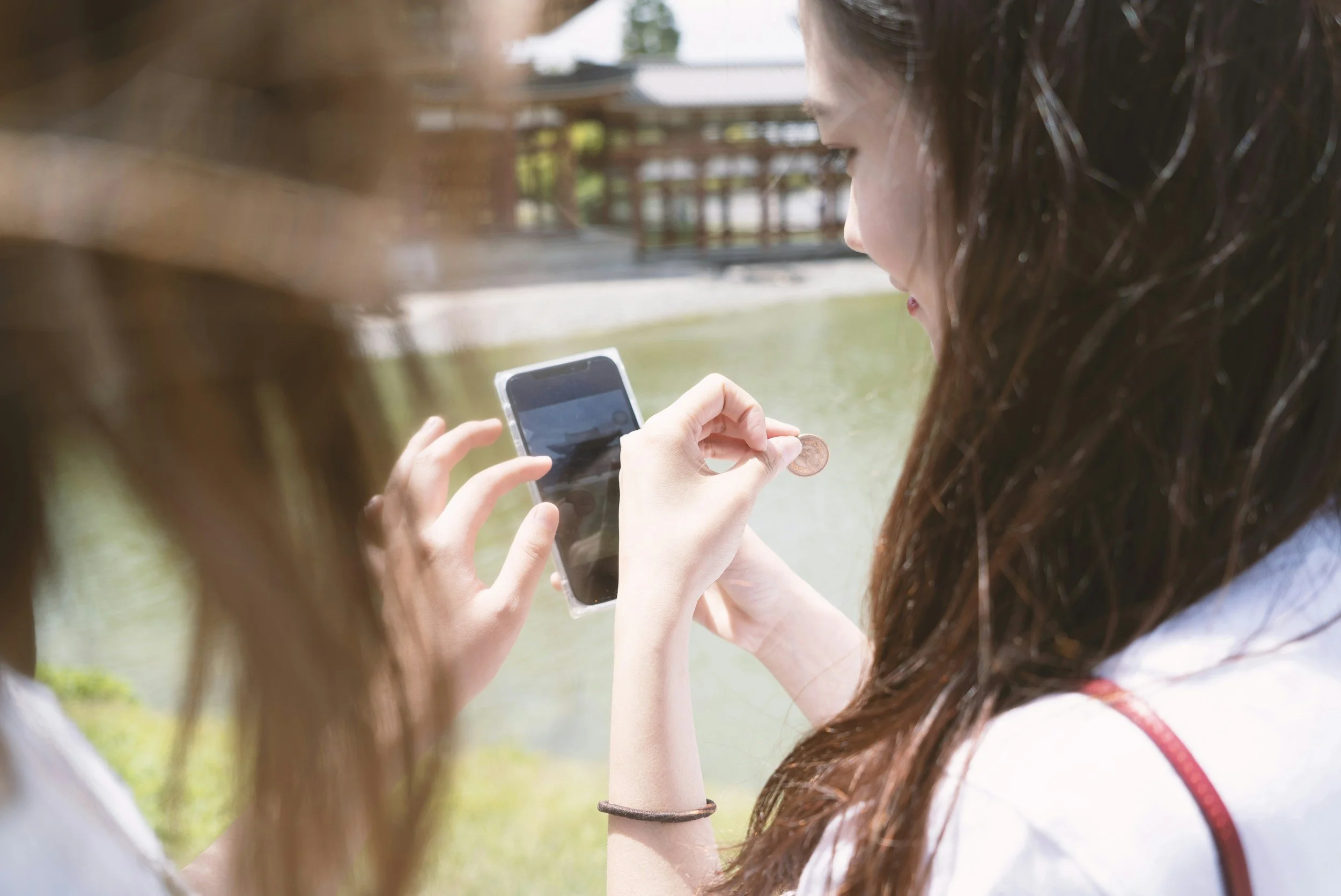 Two young women are sitting outdoors, one of them is showing her phone to the other. The woman with long curly brown hair is holding a phone, and the hand of the other woman is visible, pointing at the phone screen. The background shows a wooden structure and greenery.