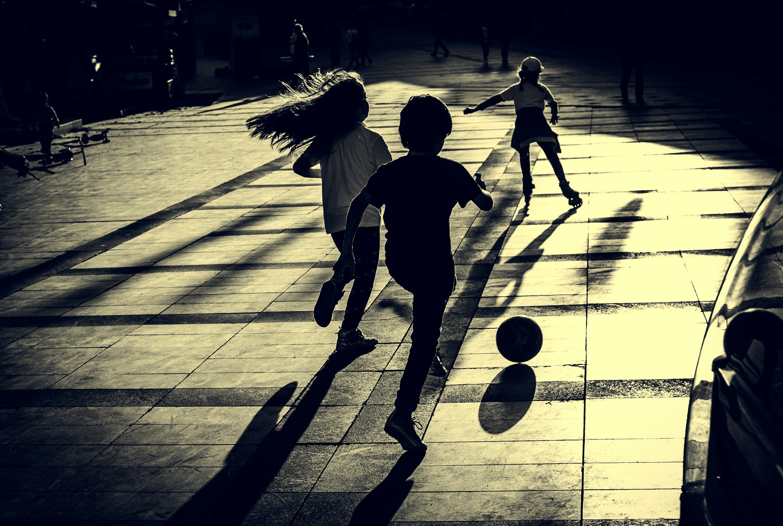 Children playing on a sunlit city street in silhouette, one is roller skating, and others are running and playing with a ball.