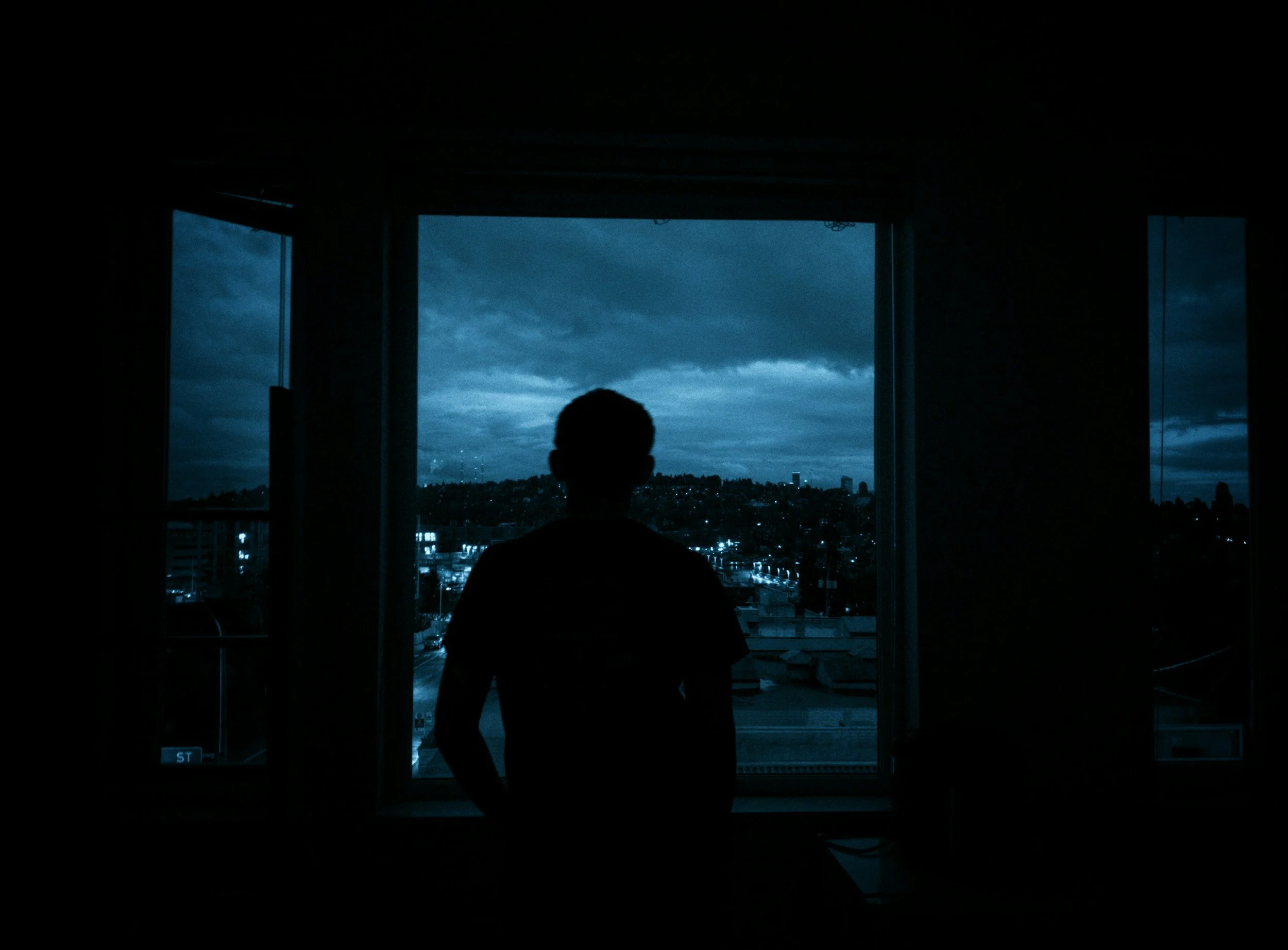 Silhouette of a person standing in front of a large window overlooking a cityscape at dusk, with dark clouds in the sky.