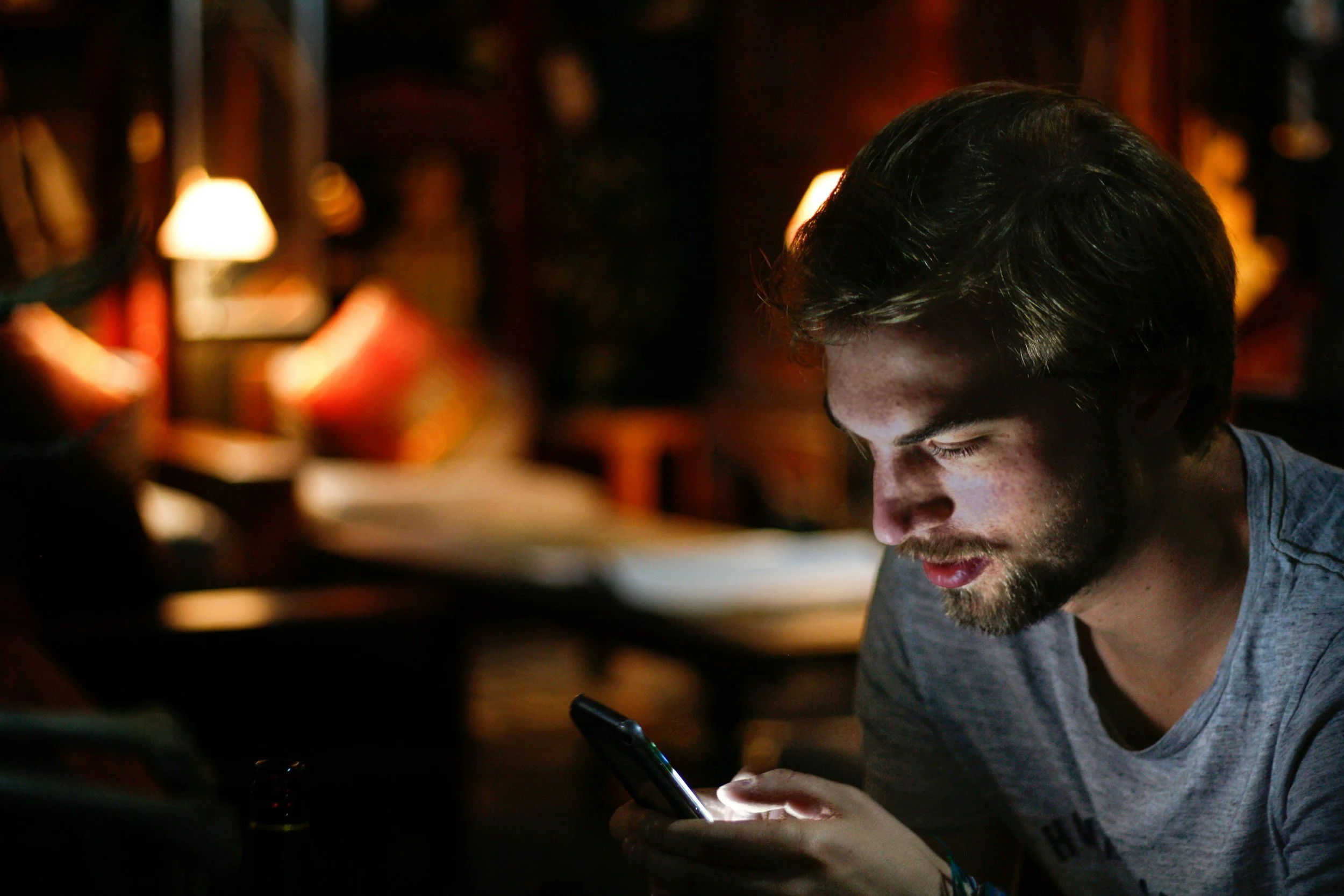 A young man with dark hair and a beard looking down at a smartphone in a dimly lit indoor setting with warm lighting and blurred background.