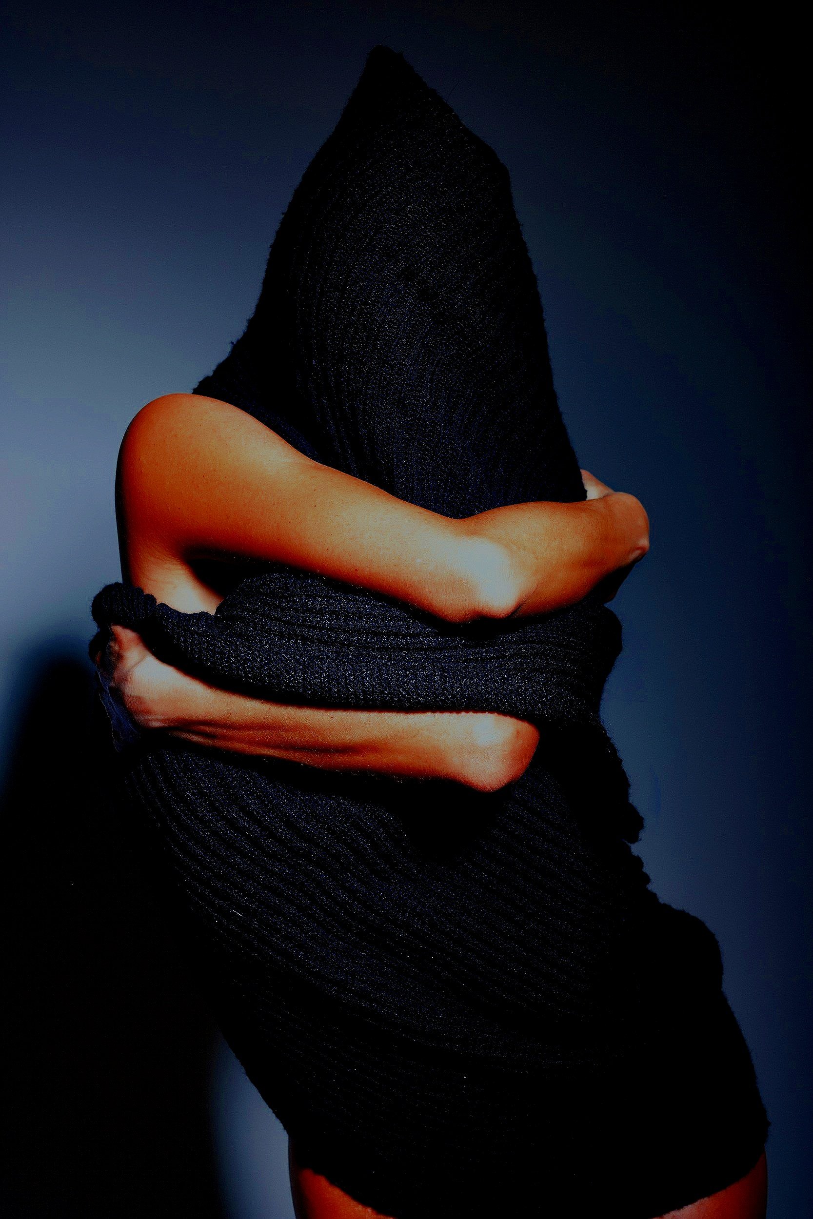 A person wearing black clothing with their arms crossed in front of their chest, with a plain dark background.