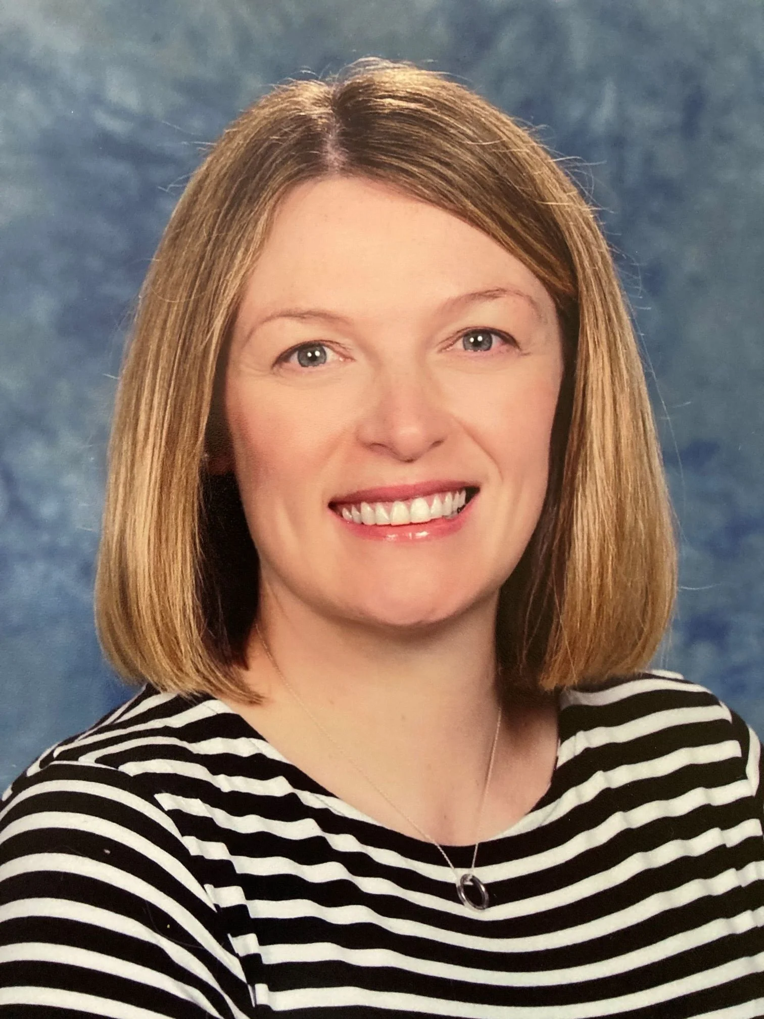A woman with shoulder-length blonde hair, smiling, wearing a black and white striped shirt and a necklace, standing against a blue textured background.