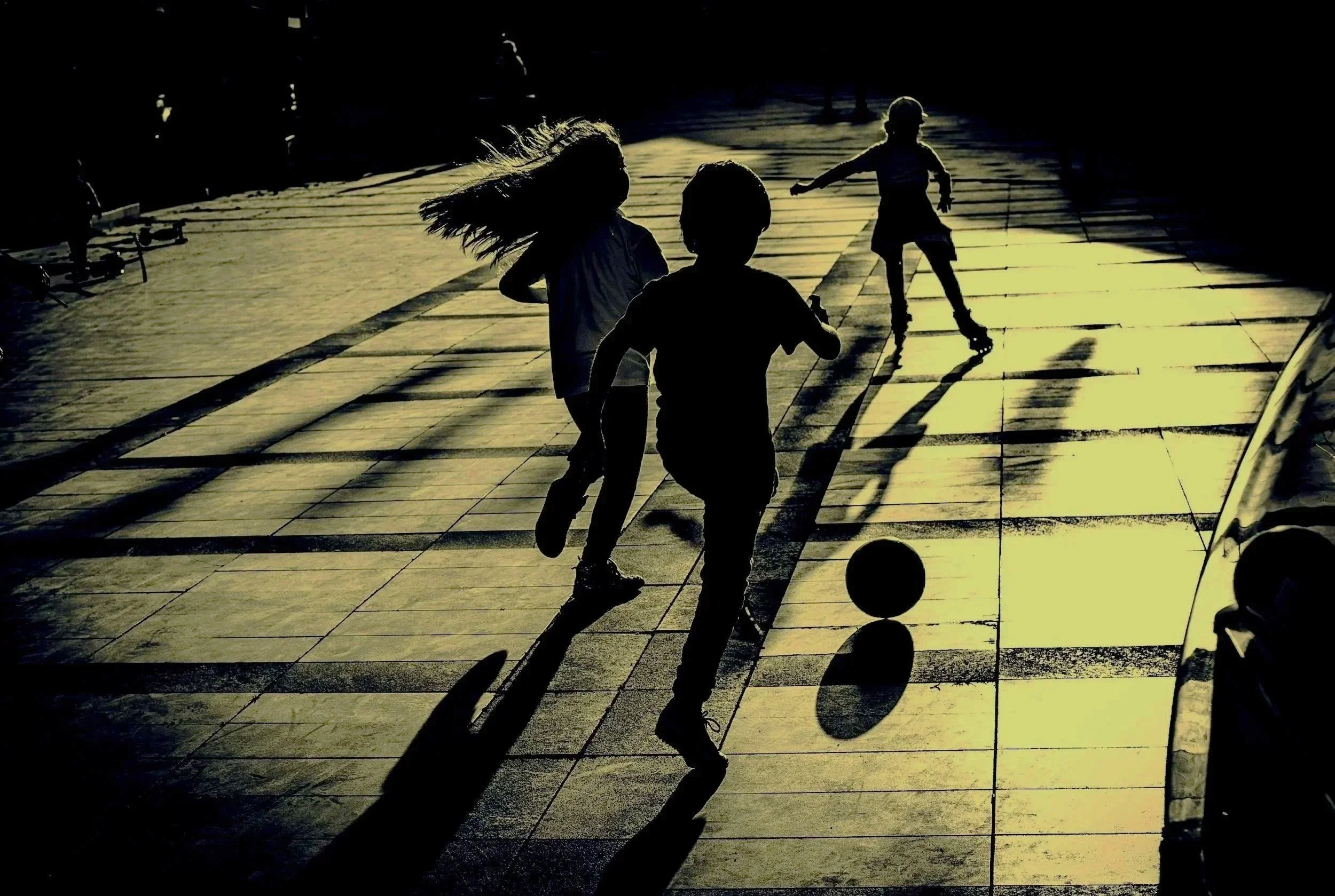 Children playing on a tiled sidewalk, with long shadows cast by the setting sun, some skating and one kicking a ball.