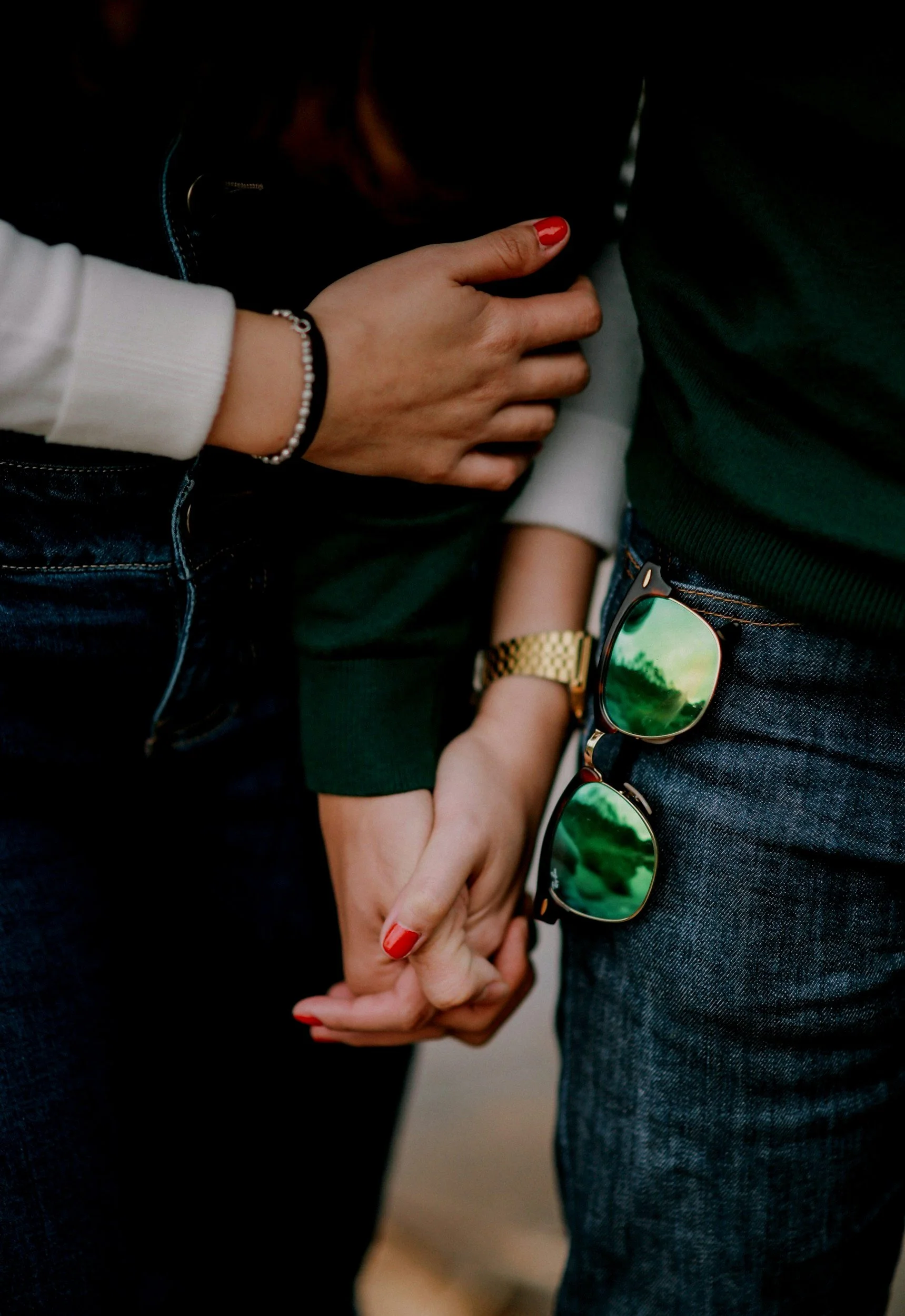 A close-up of a couple holding hands, with the woman's hand displaying red nail polish, a beaded bracelet, and a gold watch, and the man's hand partially visible, wearing jeans and a green shirt, with sunglasses hanging from his pocket.