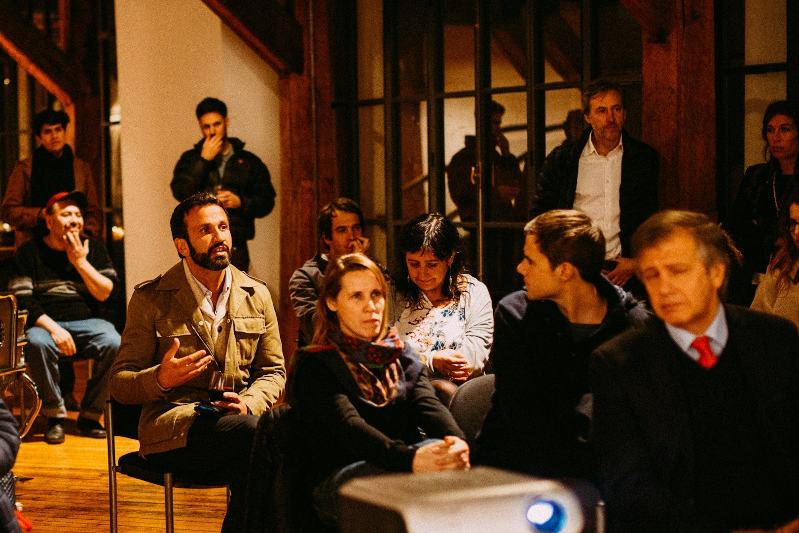 Group of people attending a meeting or event in a warmly lit room, some are seated while others stand, engaging in conversation or listening.