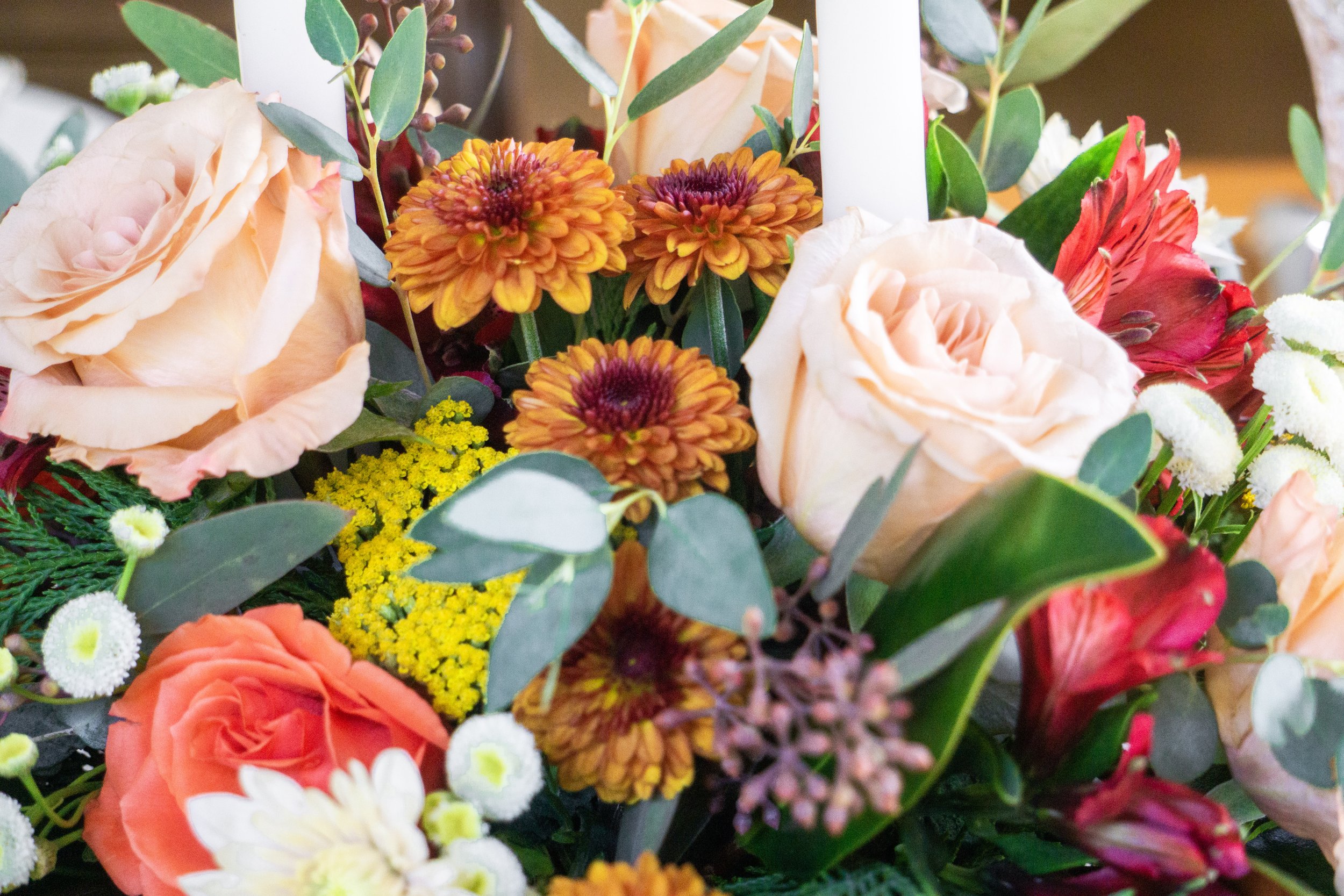 Close-up of a flower arrangement with peach roses, orange chrysanthemums, red lilies, yellow achillea, and greenery.