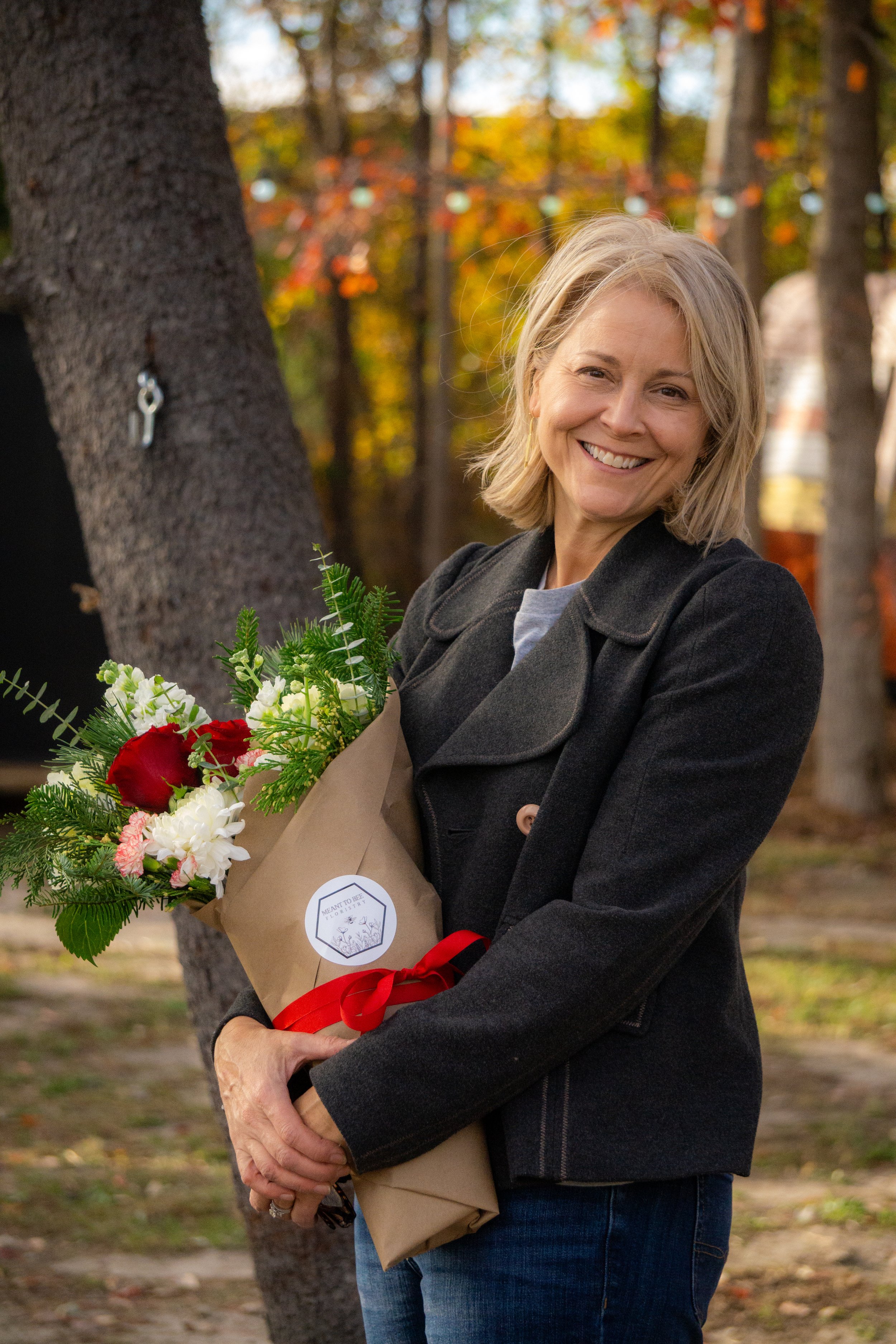 Woman smiling while holding a bouquet of flowers wrapped in brown paper with a red ribbon, standing outdoors near a tree in a park setting.