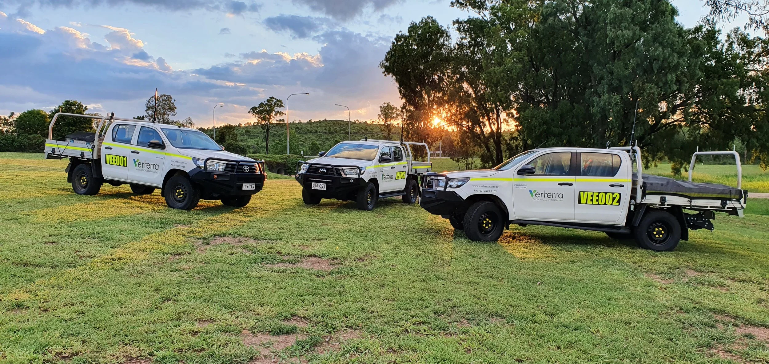 Three pickup trucks with company logos parked on a grassy field at sunset, with trees and cloudy sky in the background.