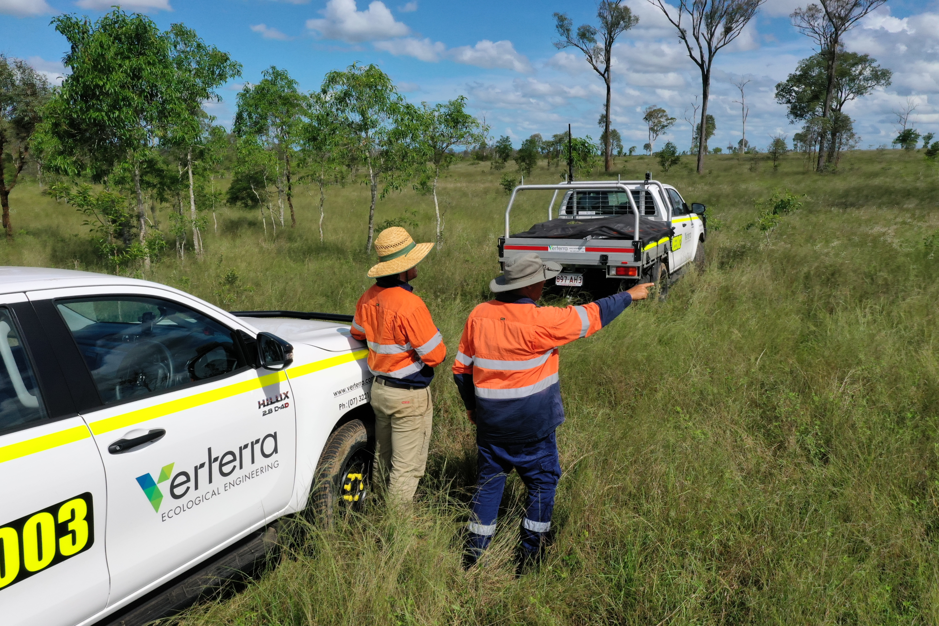 Two men in orange safety vests and hats talking to a third man in a dark uniform near a white ecological engineering vehicle with yellow stripes and a black truck bed, parked in an open grassy field with sparse trees under a bright blue sky with scattered clouds.