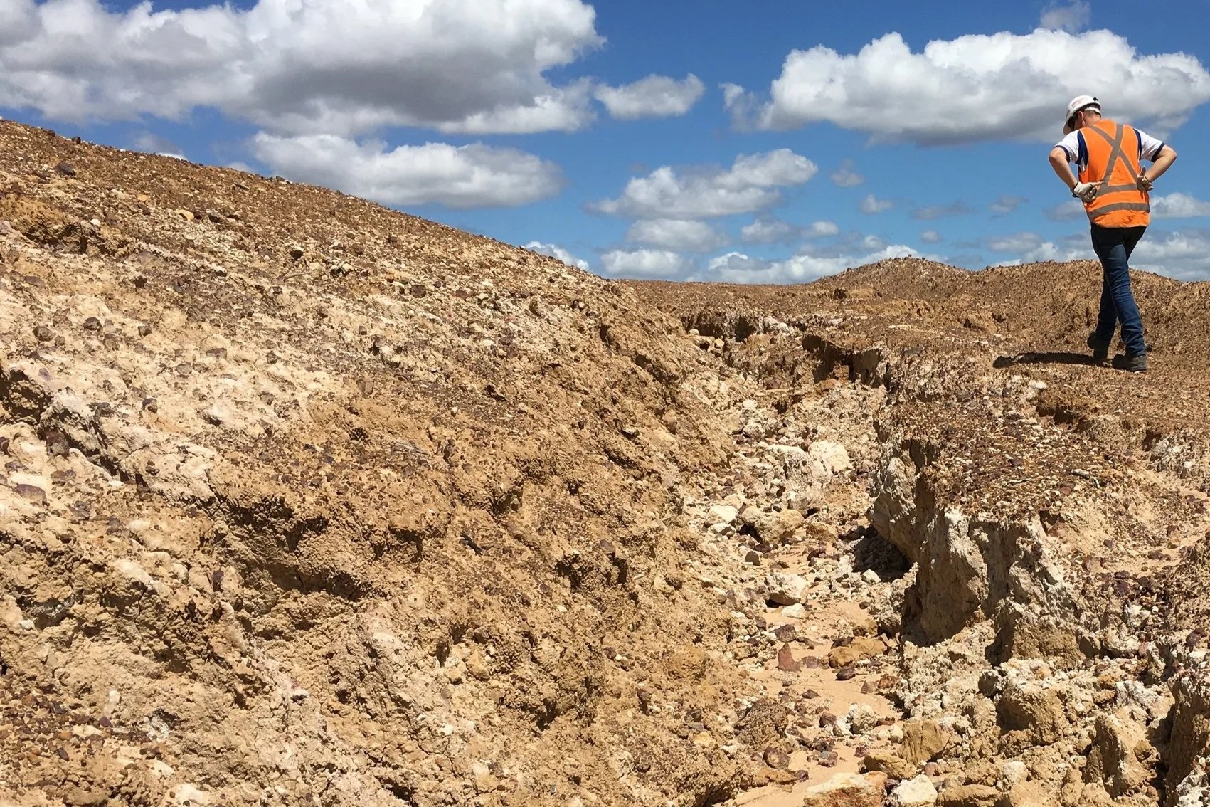 A person wearing a white hard hat, orange safety vest, blue shirt, and jeans walking along a rough, rocky terrain in a barren, desert-like landscape with mountains and a partly cloudy blue sky in the background.