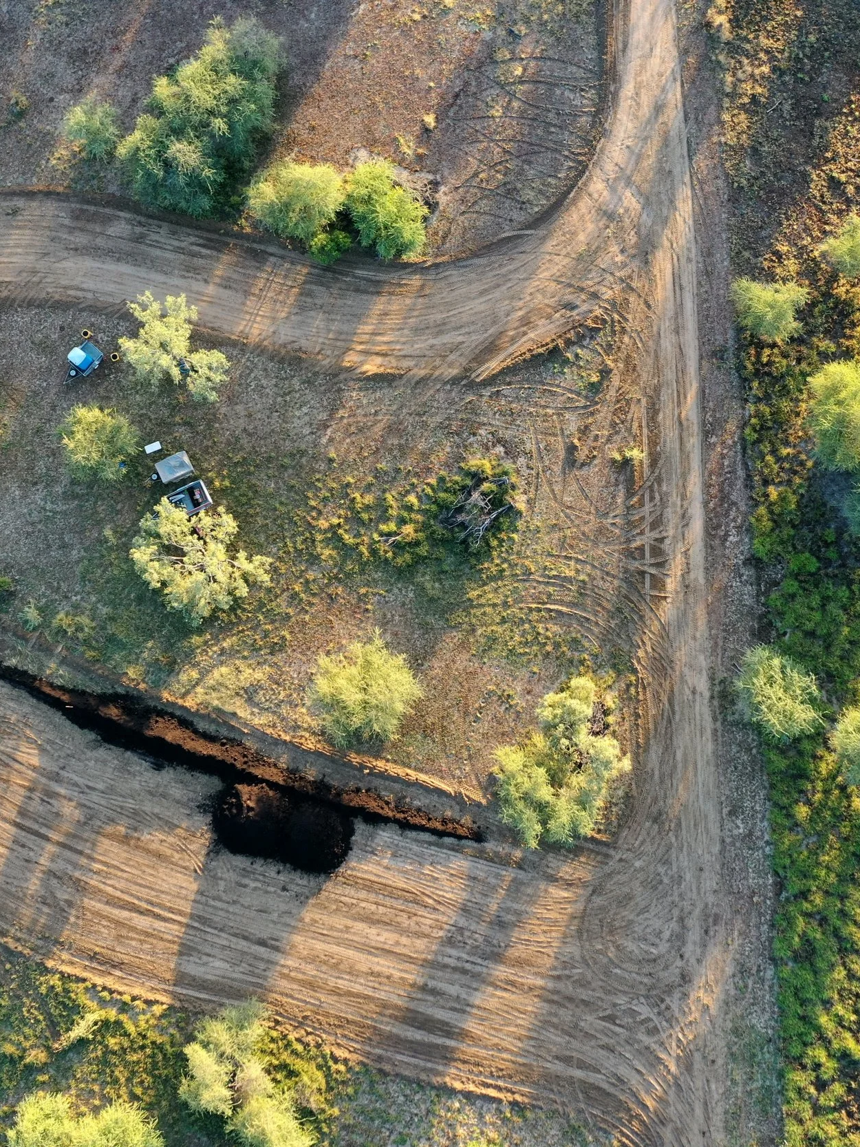 An aerial view of a construction site with a partially cleared area, trees, and dirt roads. There are some vehicles parked among trees.