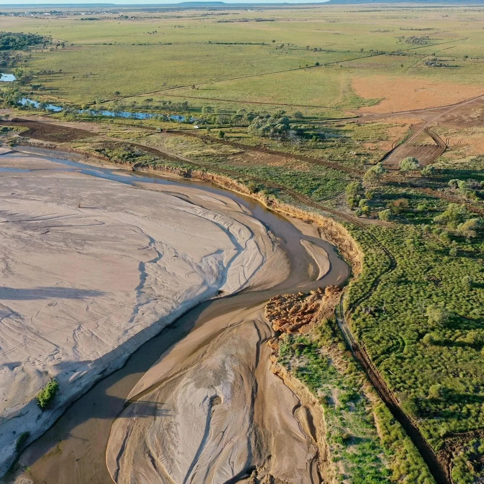 An aerial view of a landscape showing a riverbed with dry sand, patches of water, and green vegetation along its edges. Beyond the river, there are green fields, trees, and some structures with roads running through the area.