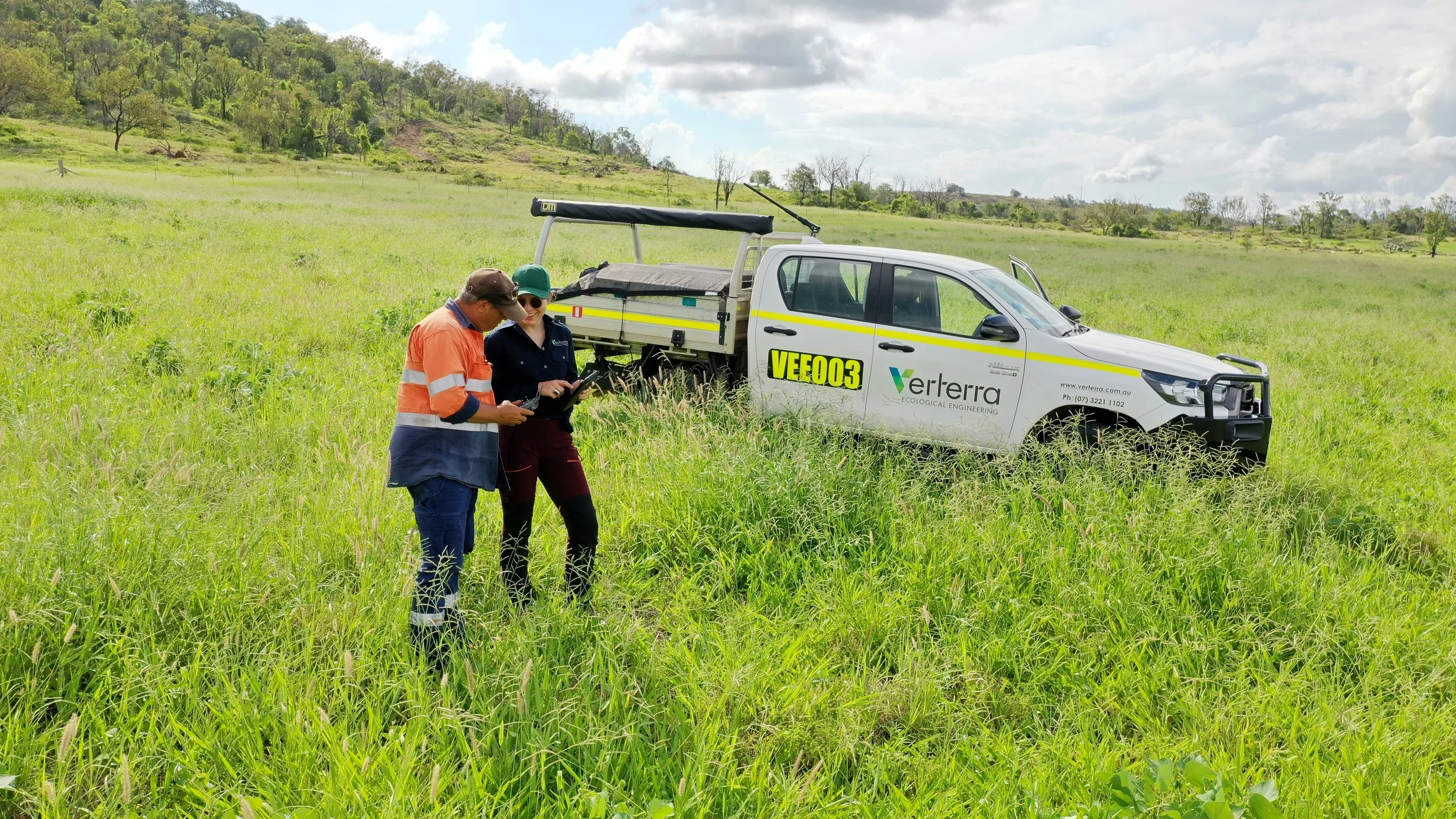 Two researchers in safety vests and hats standing in tall grass, looking at a digital tablet next to a white service vehicle with Verterra ecological engineering branding, parked in a green field with hills and trees in the background.